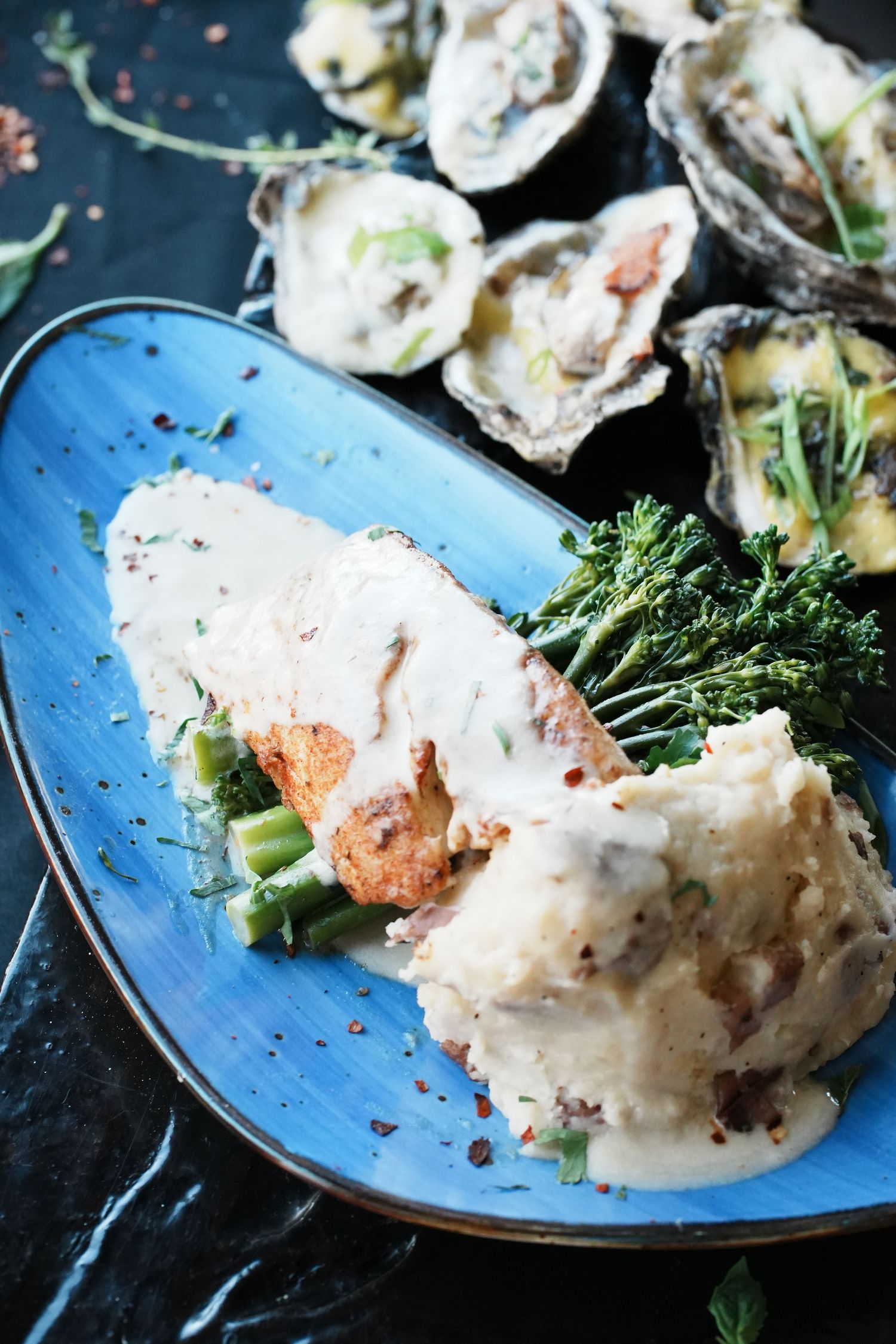 Salmon with creamy sauce and broccolini served on a bright blue plate next to fresh oysters.