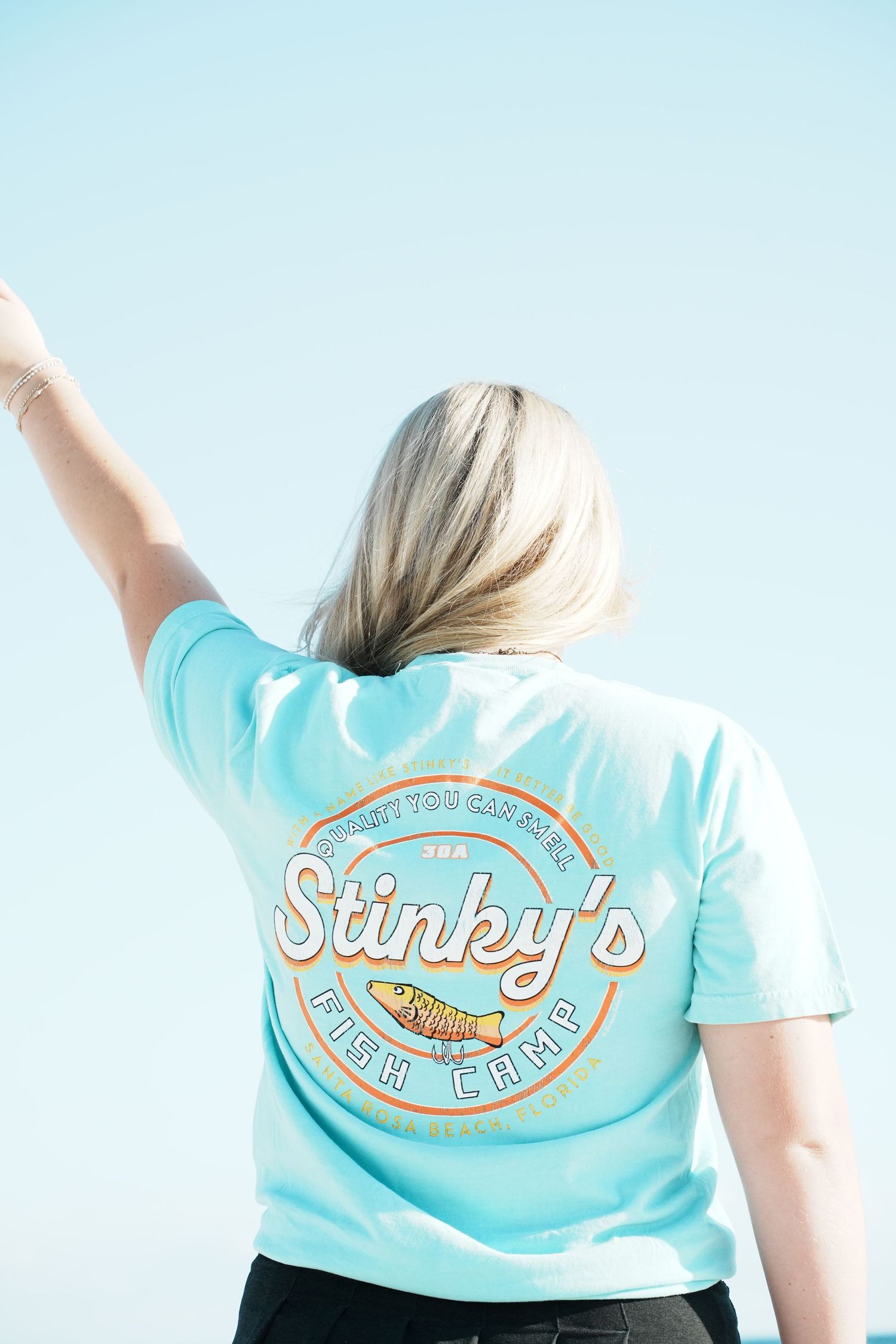 Person wearing light blue t-shirt with Sunny's Fish Camp logo stretching arms outward on wooden deck.