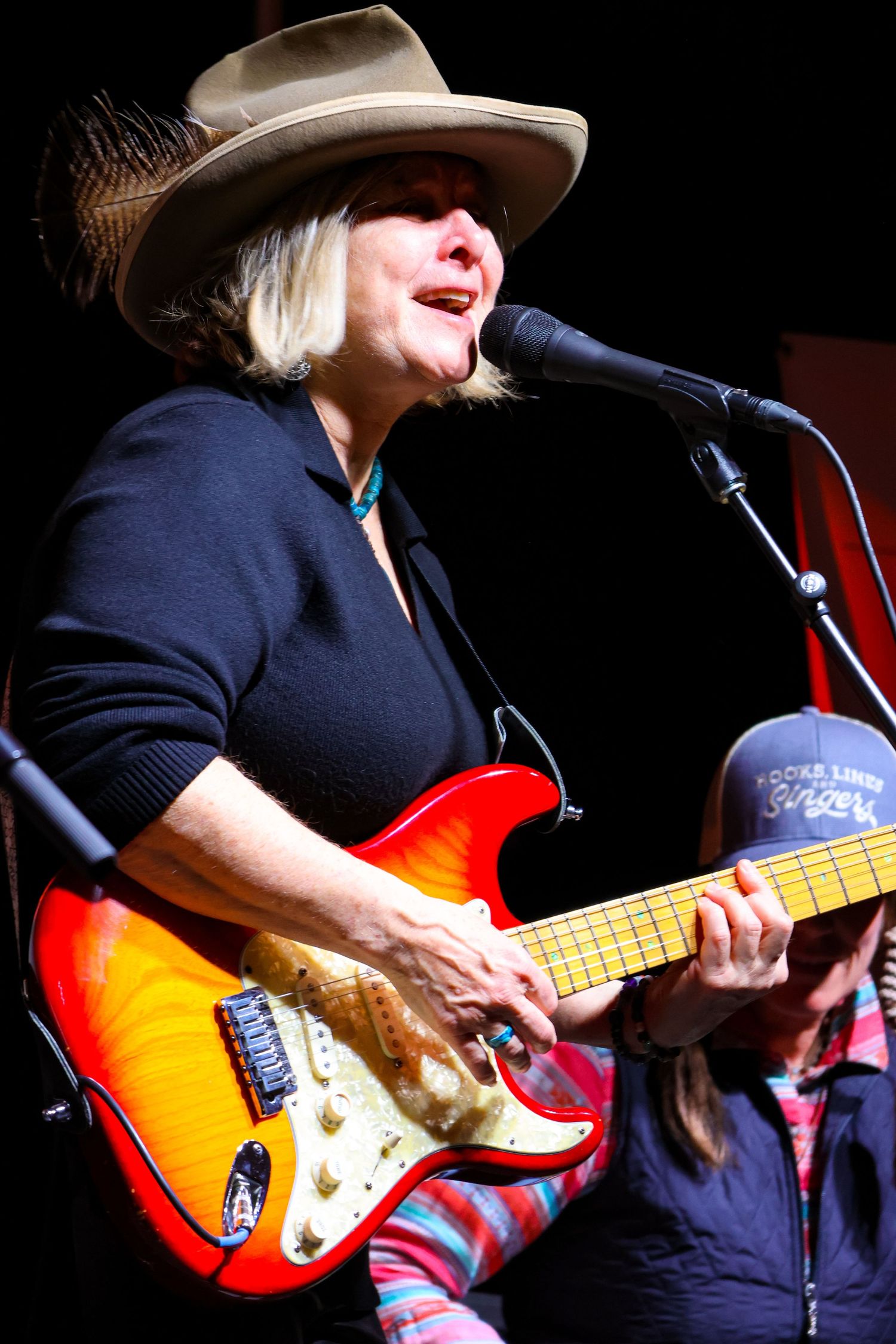 Performer playing sunburst electric guitar on dark stage with cowboy hat.