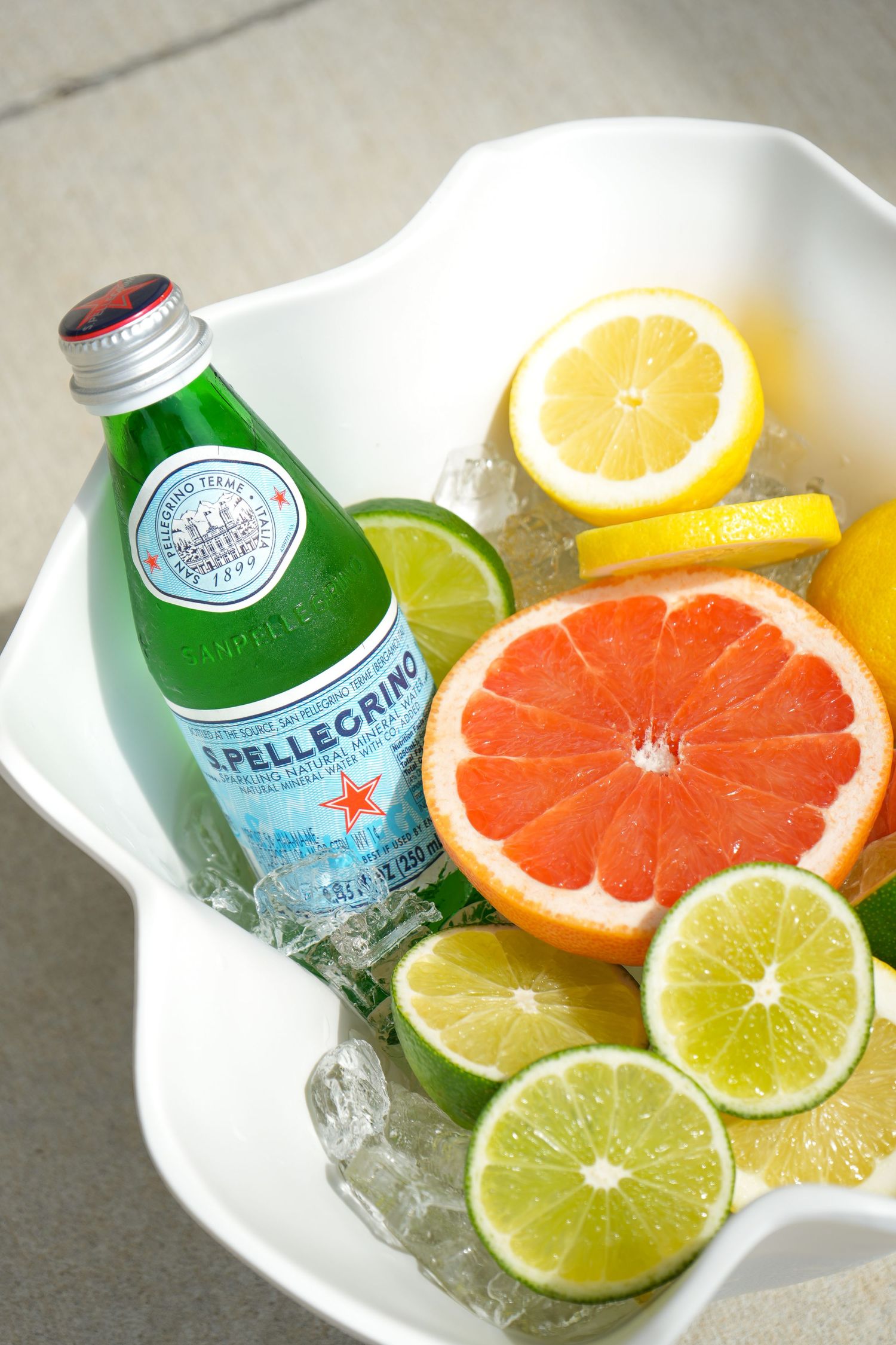 Bottle of Pellegrino sparkling water with fresh citrus slices including grapefruit, lime and lemon on ice in white bowl.