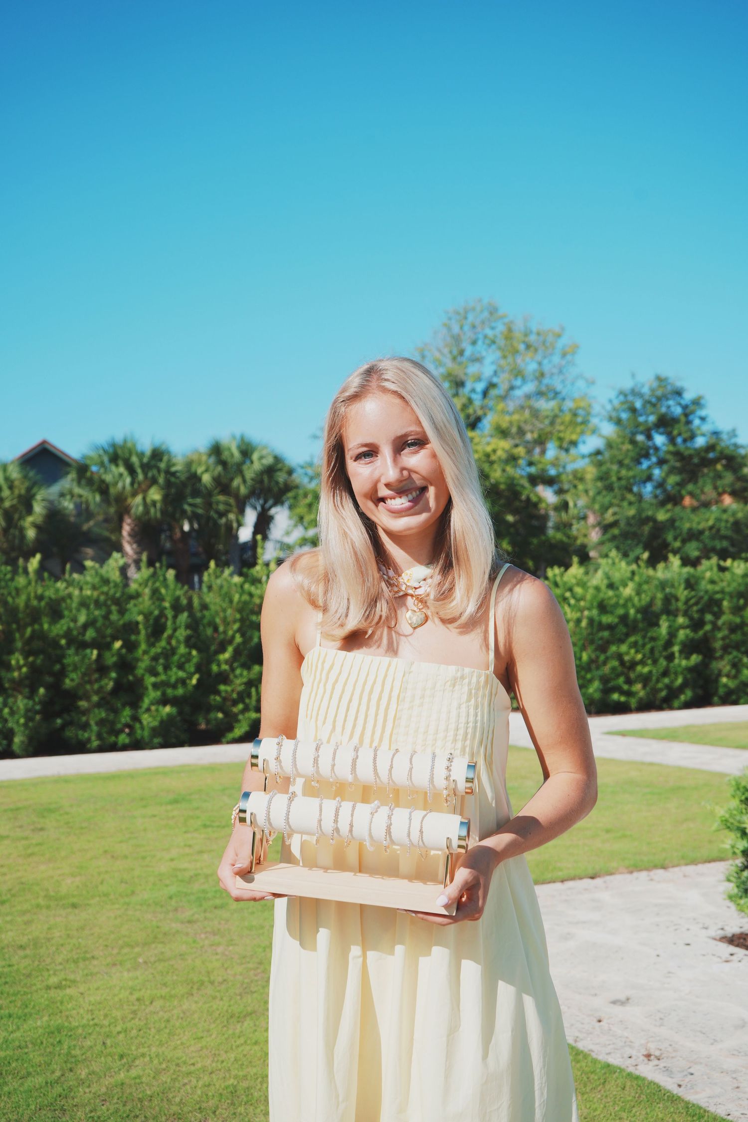Person in light colored dress stands outdoors holding a sign or letter display against blue sky and greenery backdrop.