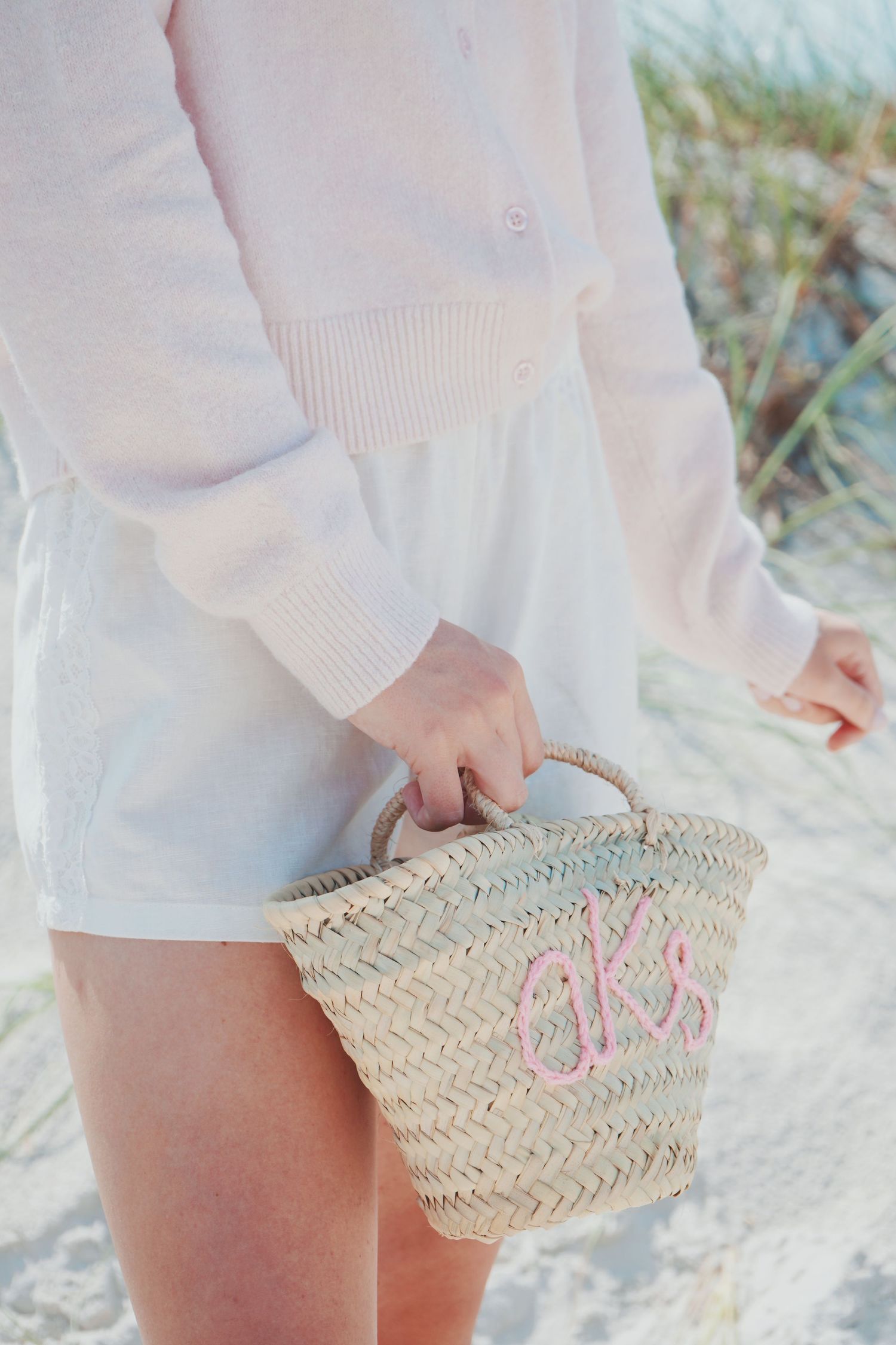 Person in a white outfit holding a woven straw beach bag with pink monogram details on sand.