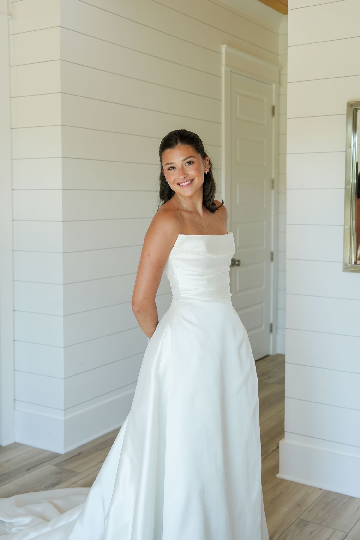 Smiling bride in a strapless white satin A-line wedding gown posing in a bright room with shiplap walls.