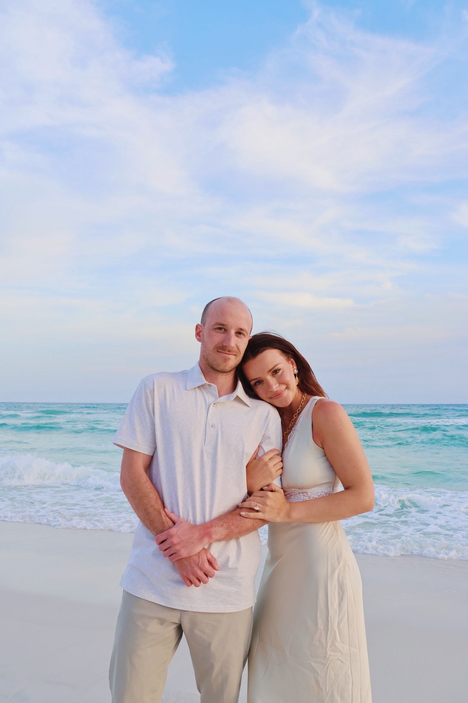 A couple in coordinated white and beige outfits embraces on a beautiful turquoise beach during a romantic photoshoot.