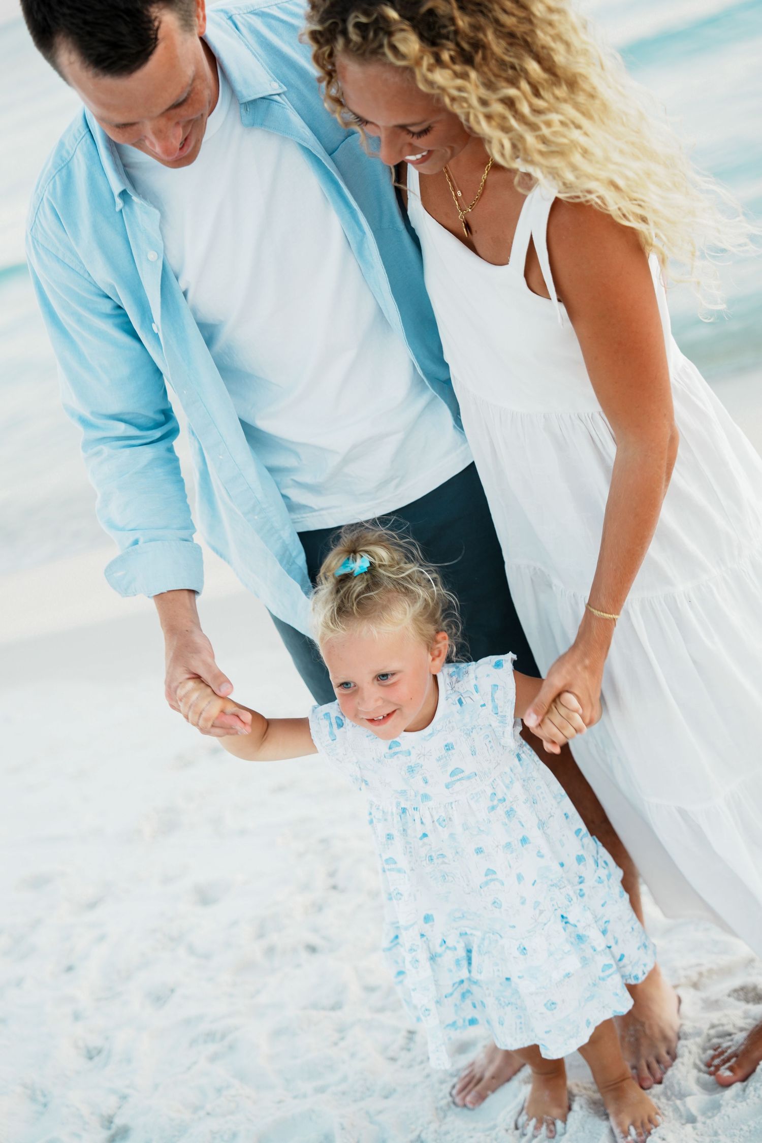 Family in coordinating blue and white outfits holding hands and smiling together on a sandy beach.