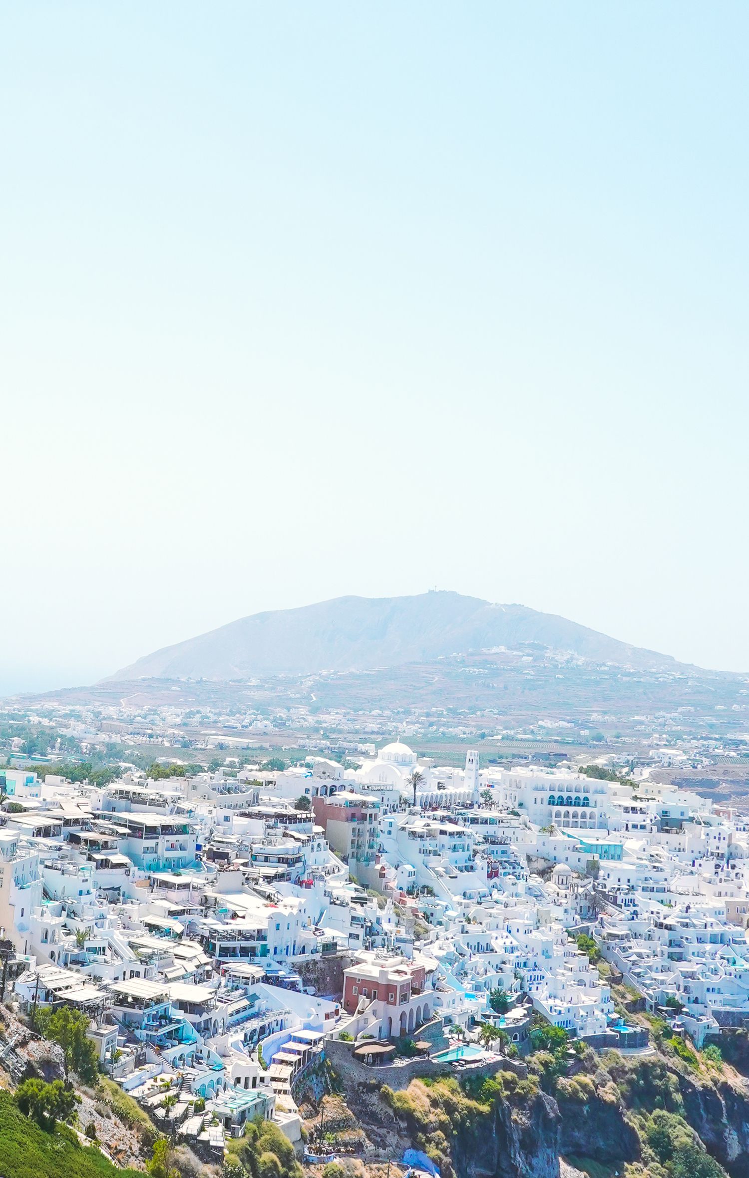 Panoramic view of Santorini with white buildings cascading down the hillside against blue sky.