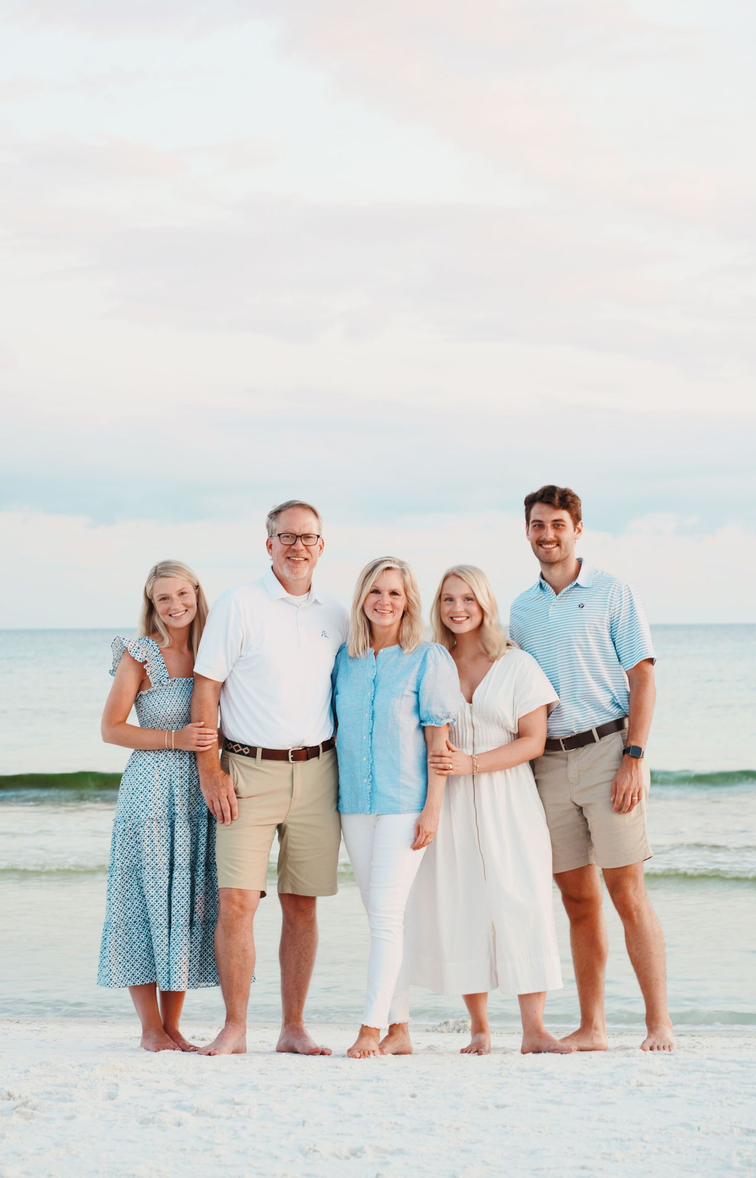 A group poses together on a white sandy beach wearing coordinated white, beige and light blue beach attire.