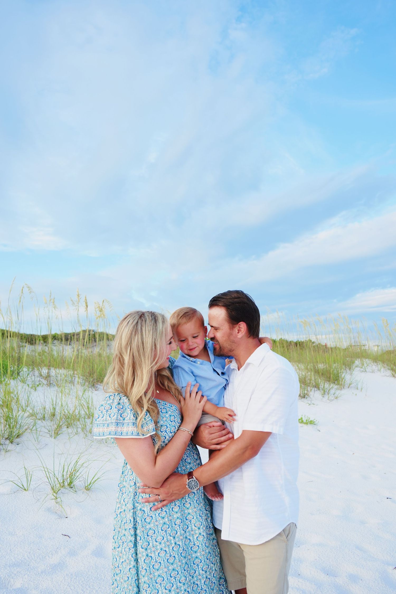 A loving family moment captured on a scenic beach with sea oats and blue skies.