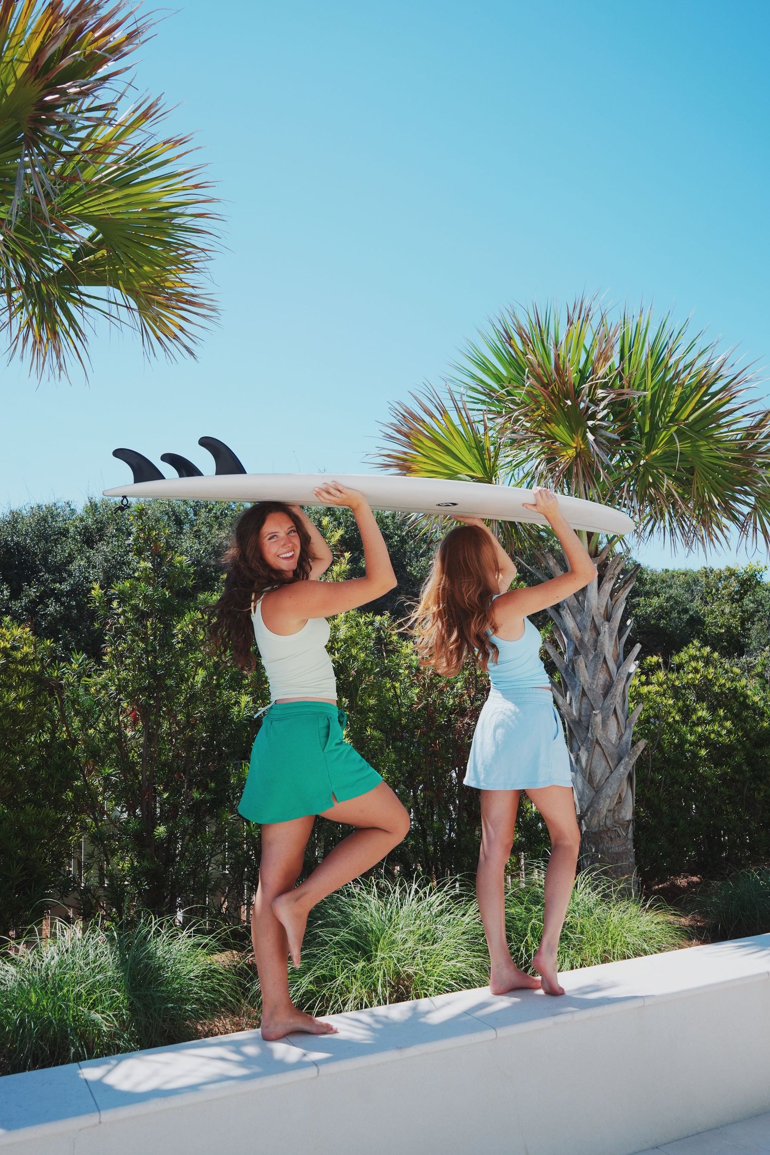 Two surfers in summer dresses holding a surfboard overhead against palm trees and blue sky on a sunny day.