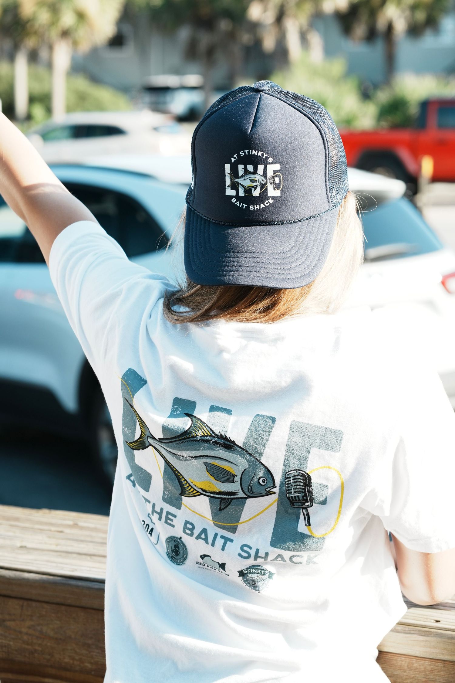 Person wearing a white t-shirt with fish design and grey LSE baseball cap viewed from behind.
