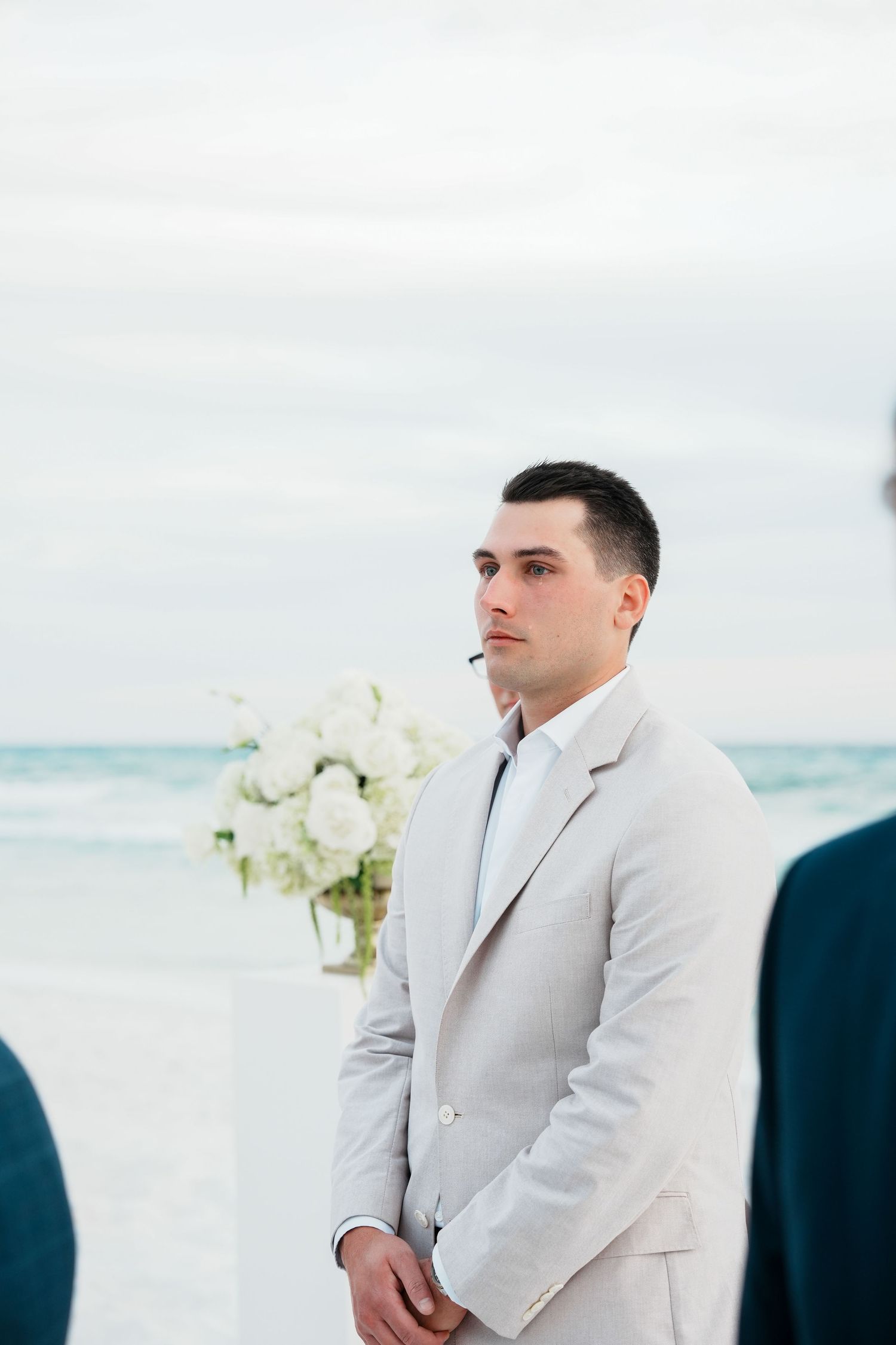 A wedding ceremony takes place on a serene beach with white flowers and light gray attire against the ocean backdrop.
