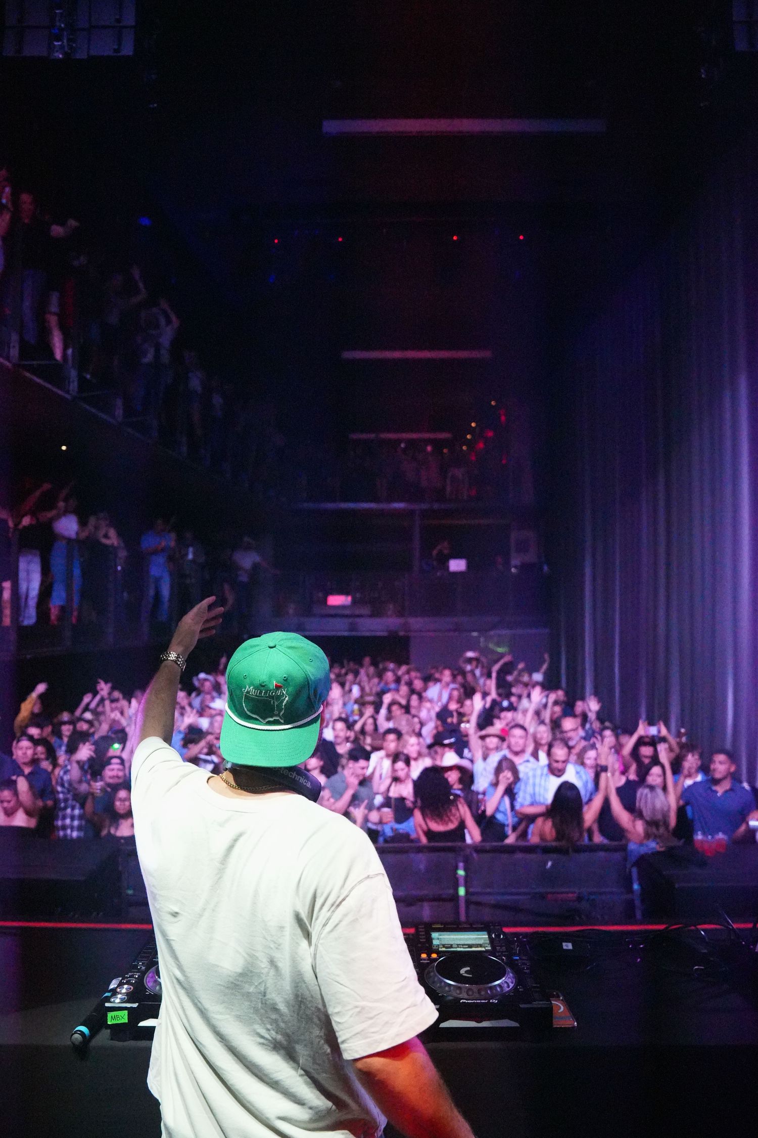 DJ performs on stage at a nightclub with large crowd visible in background under purple lighting.