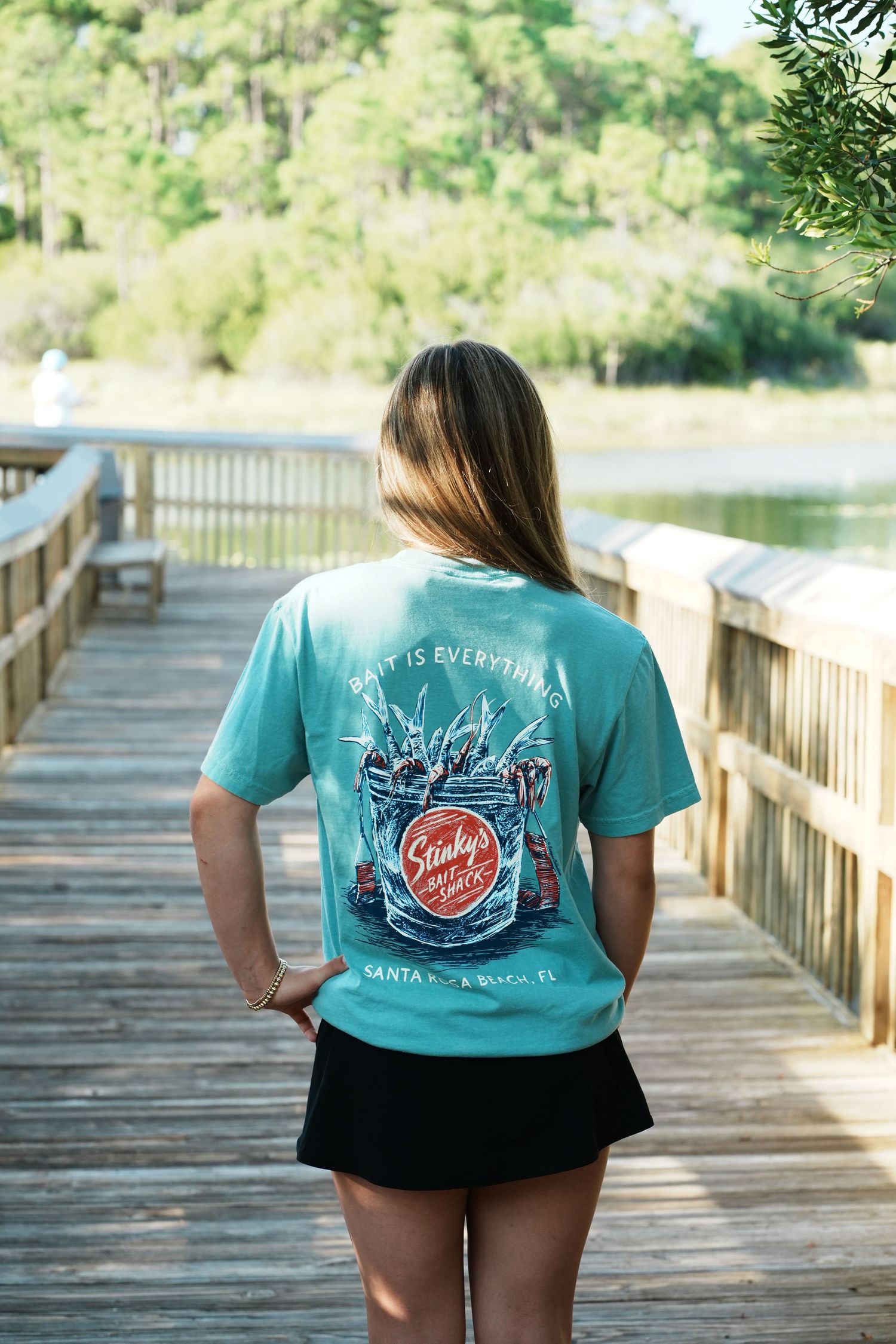 Person wearing turquoise tshirt with coastal design standing on wooden boardwalk over water.