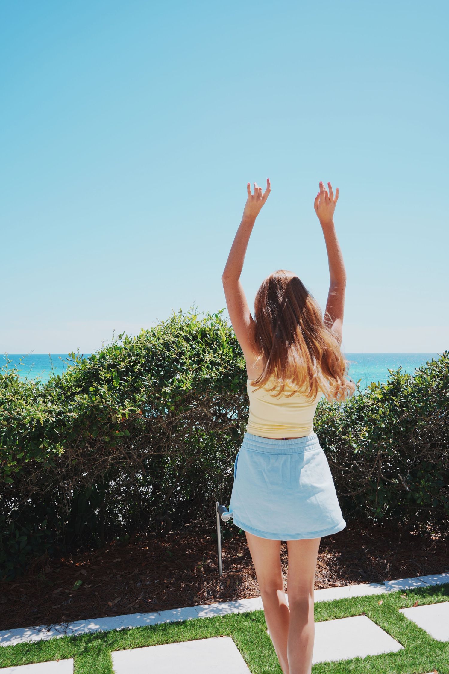 Woman in yellow top and light skirt raises arms joyfully against blue ocean and sky backdrop.
