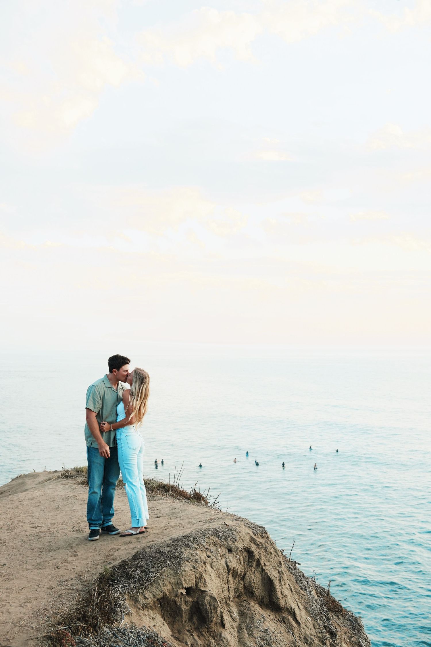 Couple embraces romantically on a scenic cliff overlooking turquoise ocean waters during sunset.