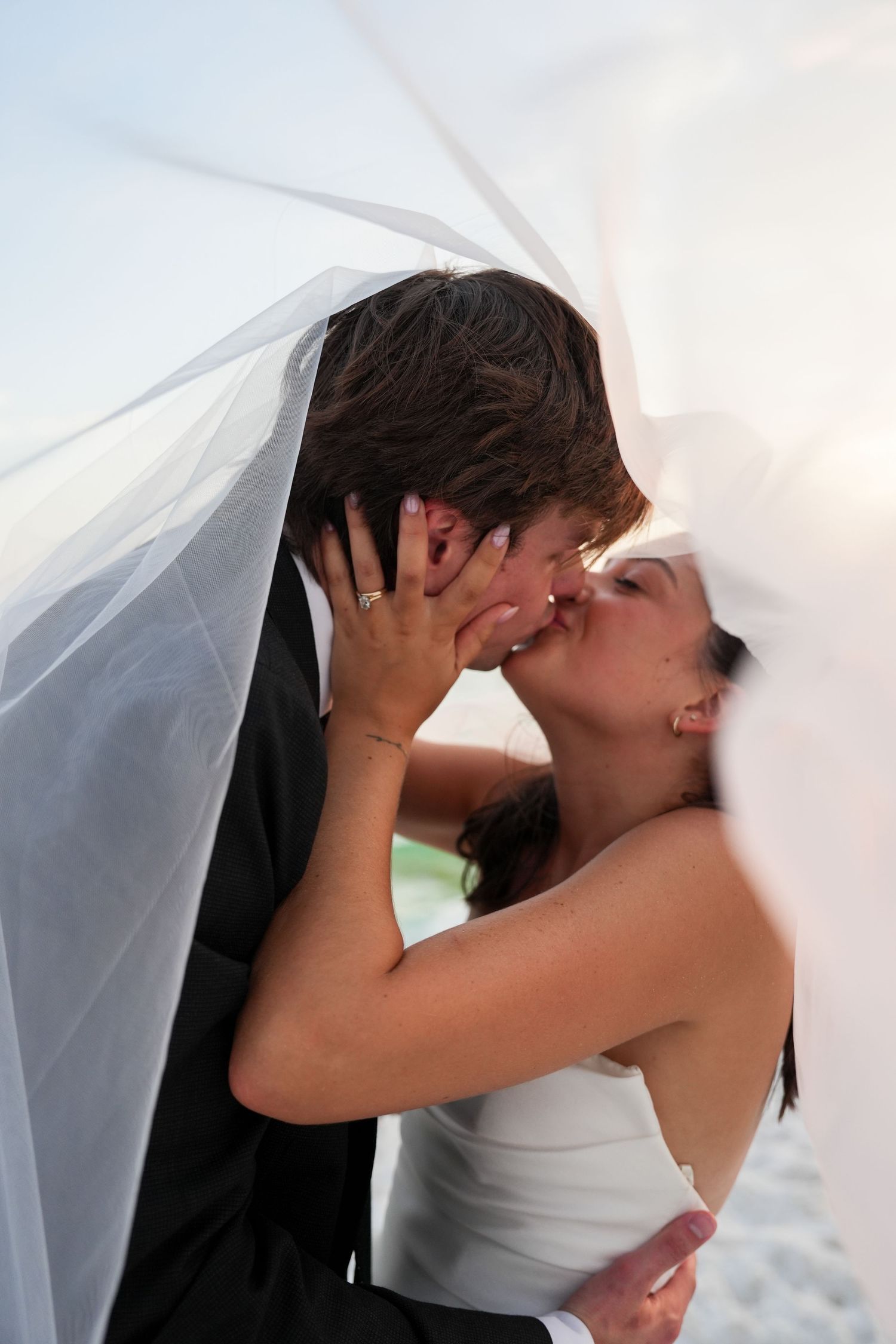 Bride and groom sharing a romantic kiss under a flowing white wedding veil outdoors.