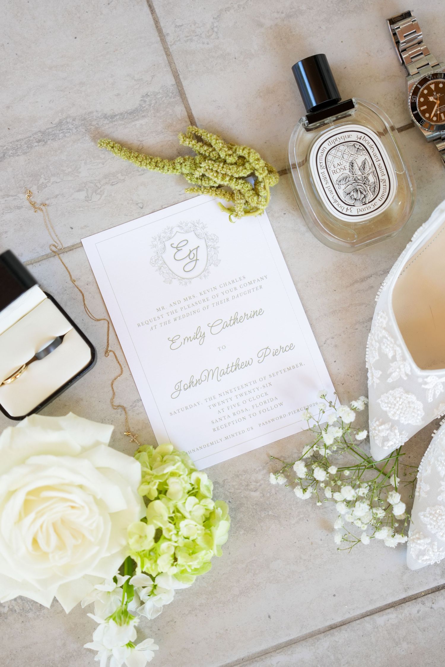 Elegant wedding flat lay with invitation, white lace heels, roses, perfume, and a watch on marble.
