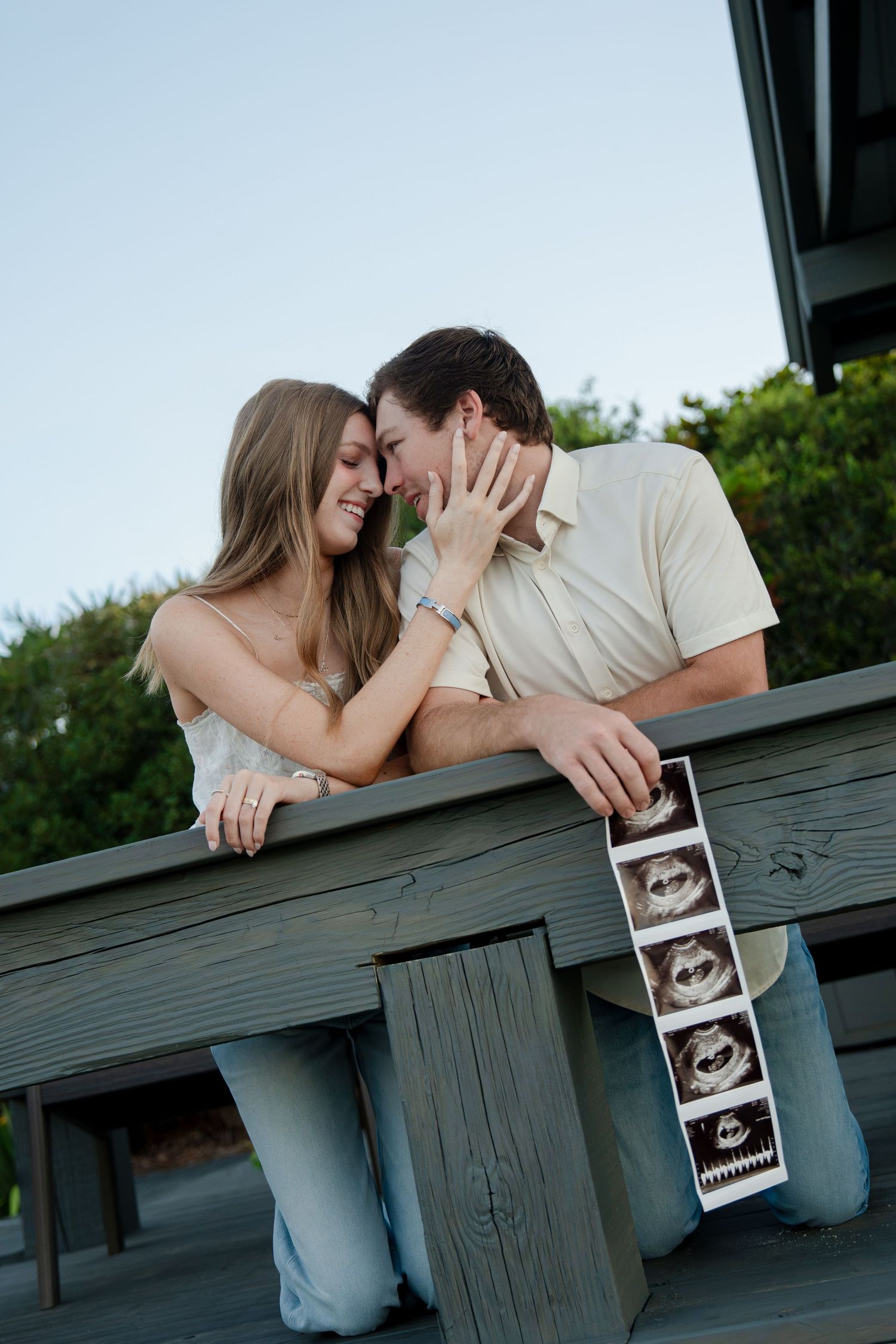 Happy couple sharing a tender moment on a deck, holding ultrasound photos announcing their pregnancy.