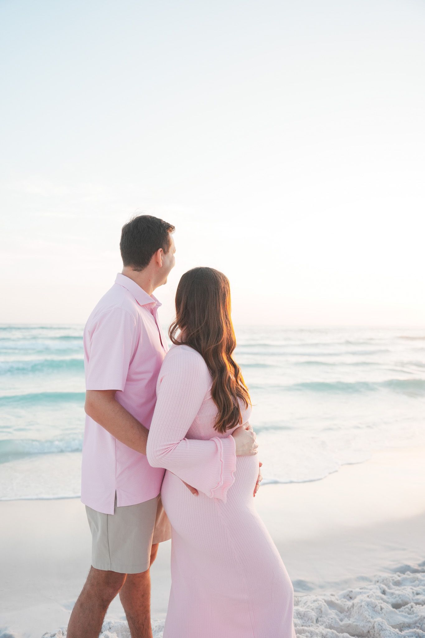 A couple in pink and white outfits embrace while walking along a serene beach at sunset.