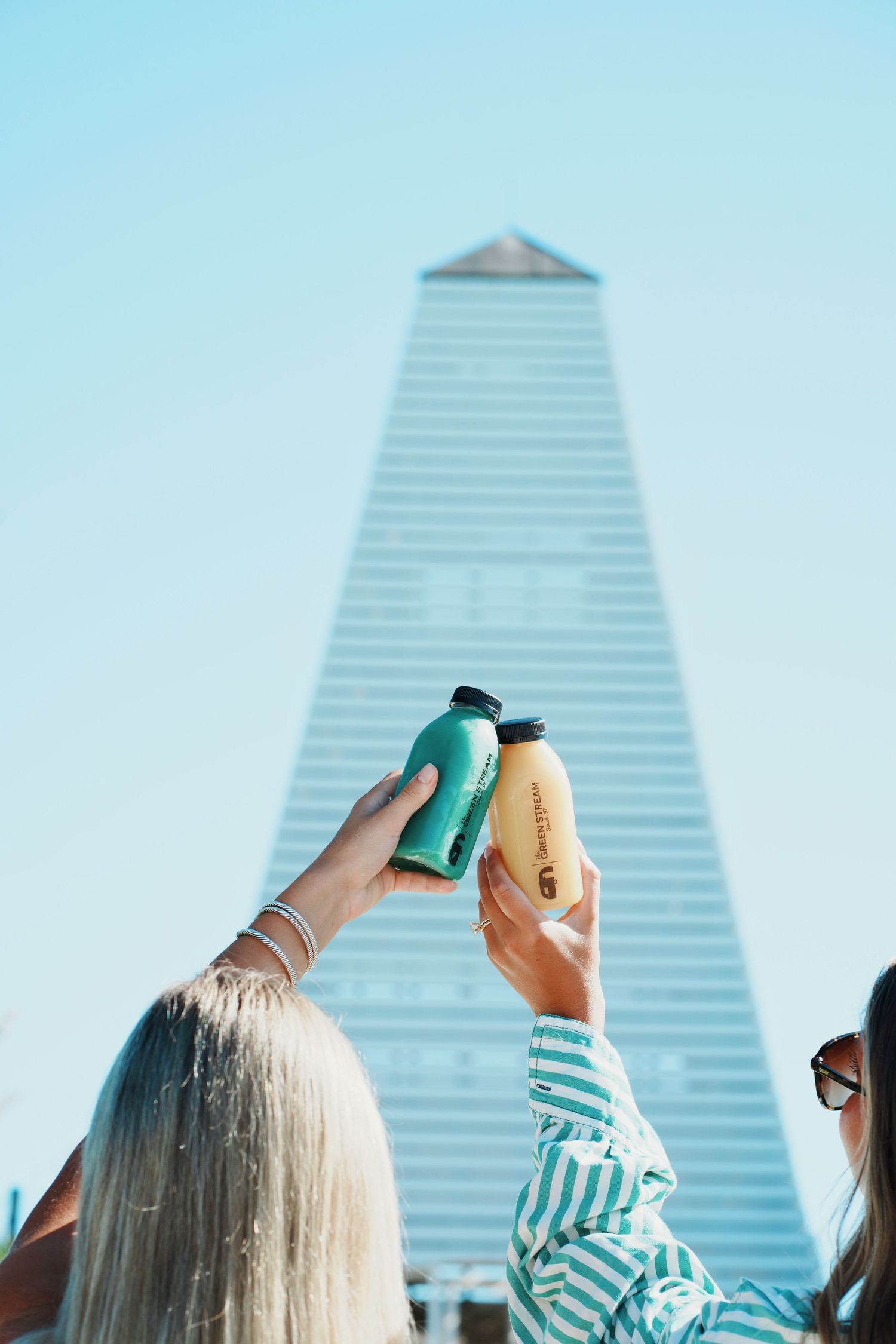Hands holding colorful drink containers against blue sky with modern building in background.