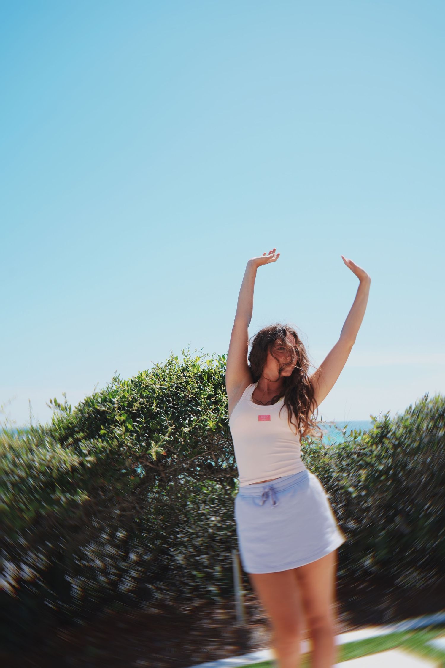 Person in white top and light skirt stretching arms upward against bright blue sky.