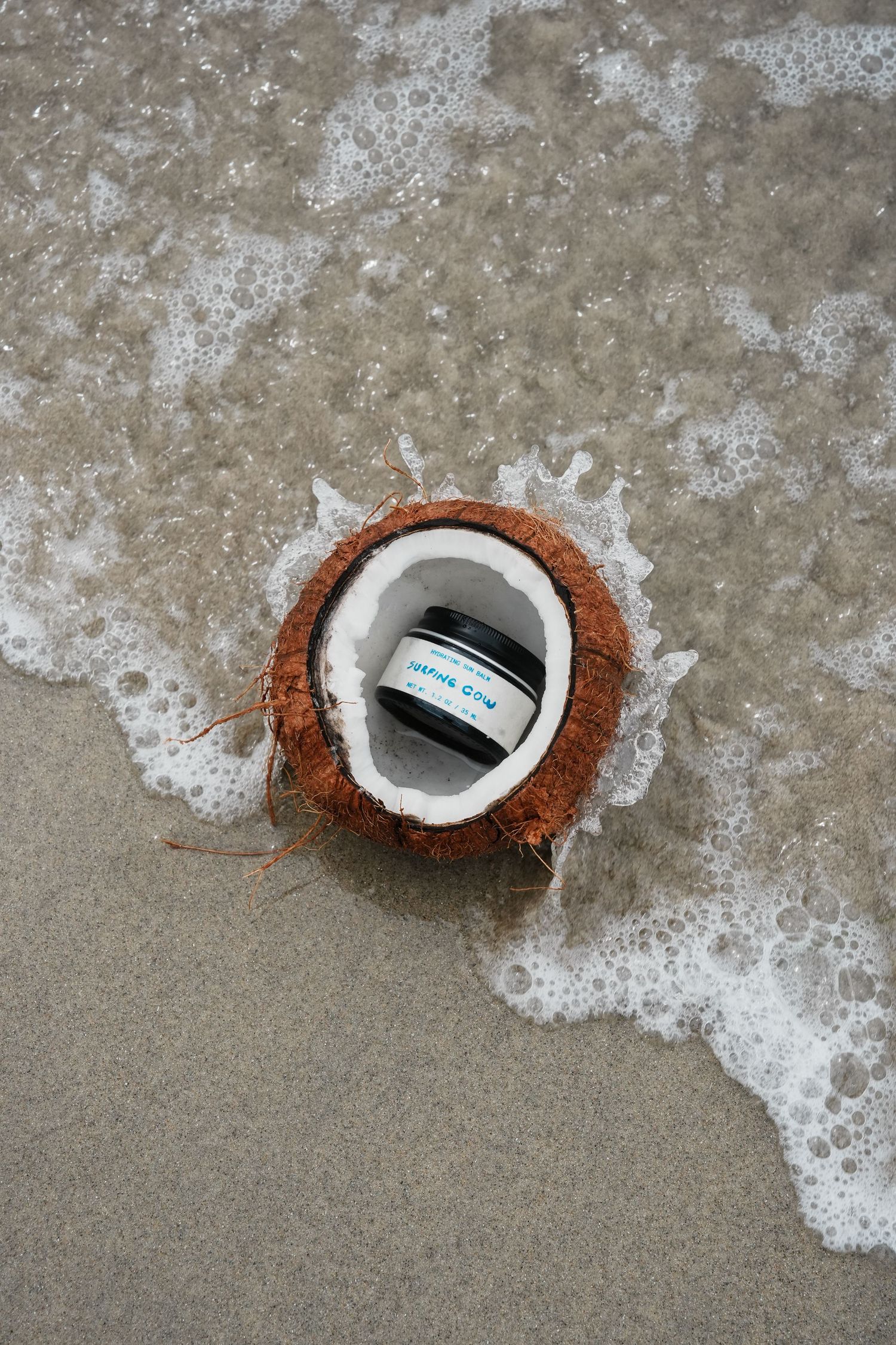 Coconut shell with product container inside being washed by ocean wave on sandy beach.