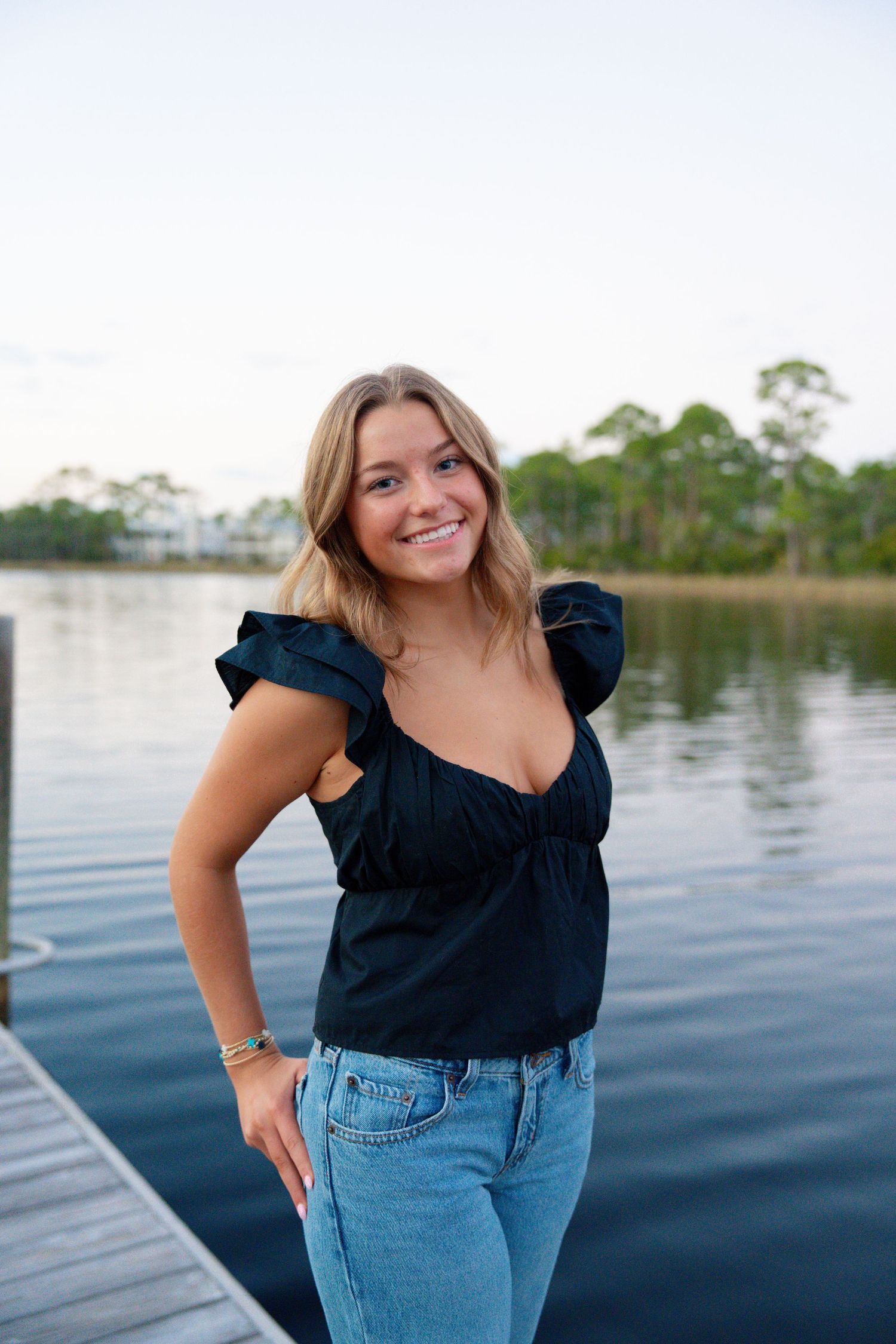 Woman in navy ruffled top and jeans poses by waterfront dock on sunny day.