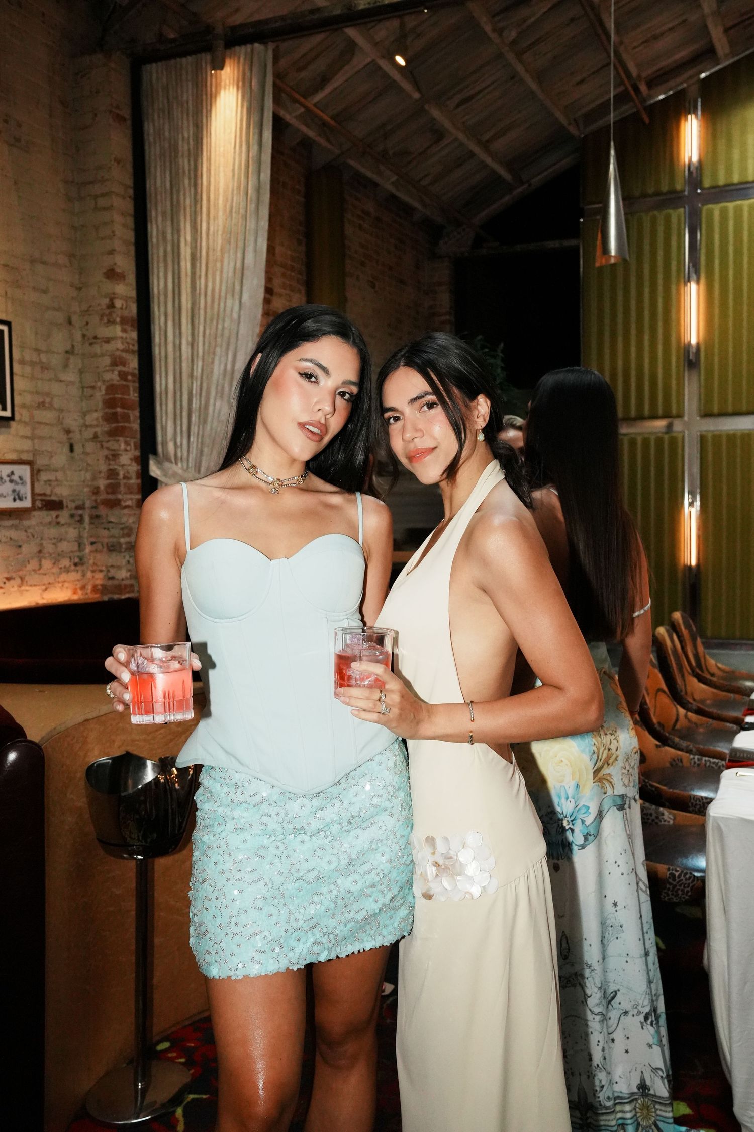 Two stylish women in elegant dresses holding cocktails at an upscale indoor event venue.
