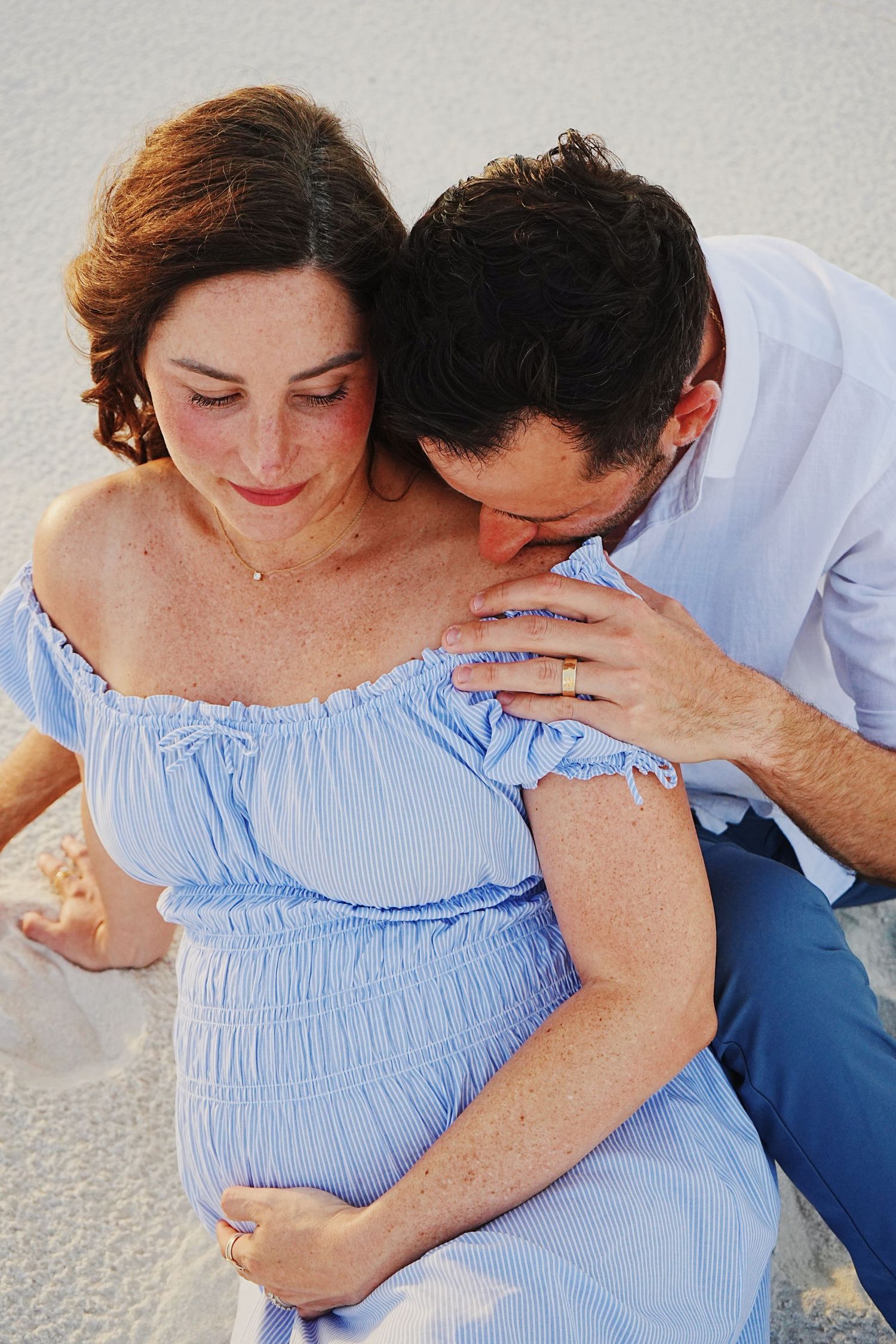 Couple sitting together in intimate maternity photo session wearing light blue dress and white shirt against neutral background.