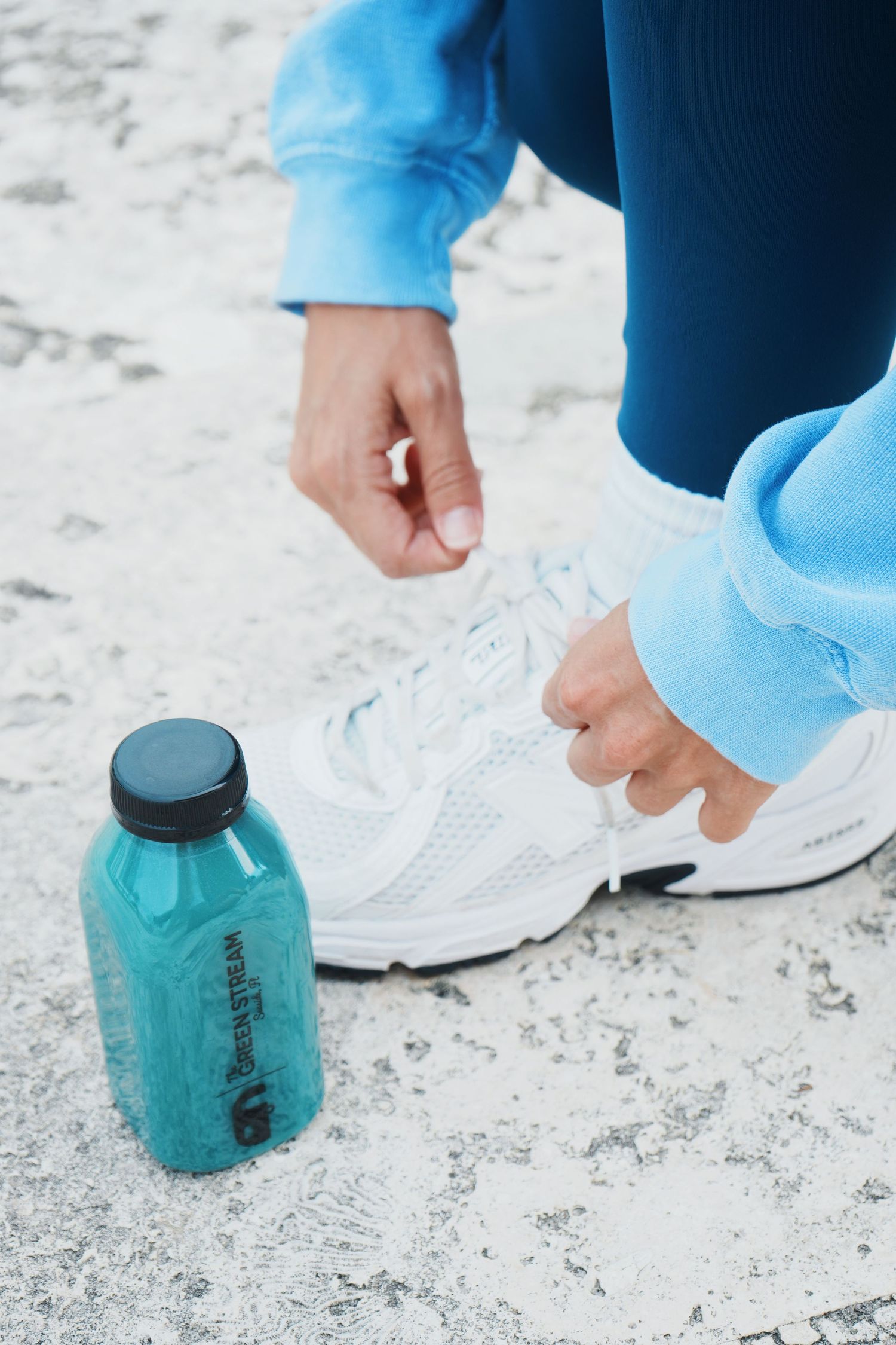 Person in blue pants tying white athletic shoes on a textured floor with a teal water bottle nearby.