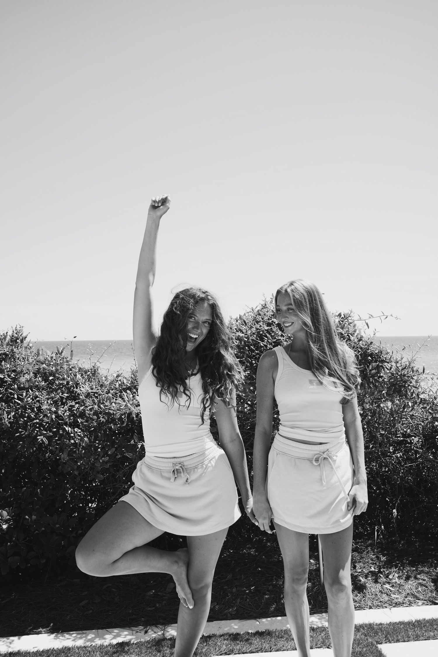 Two people in matching white outfits posing playfully against greenery in black and white.
