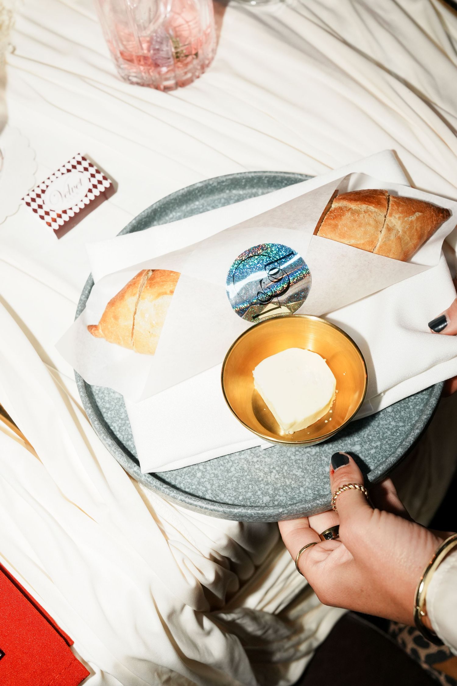 A hand holding a tray with bread rolls, butter in a gold bowl, and a decorative sticker on white paper.