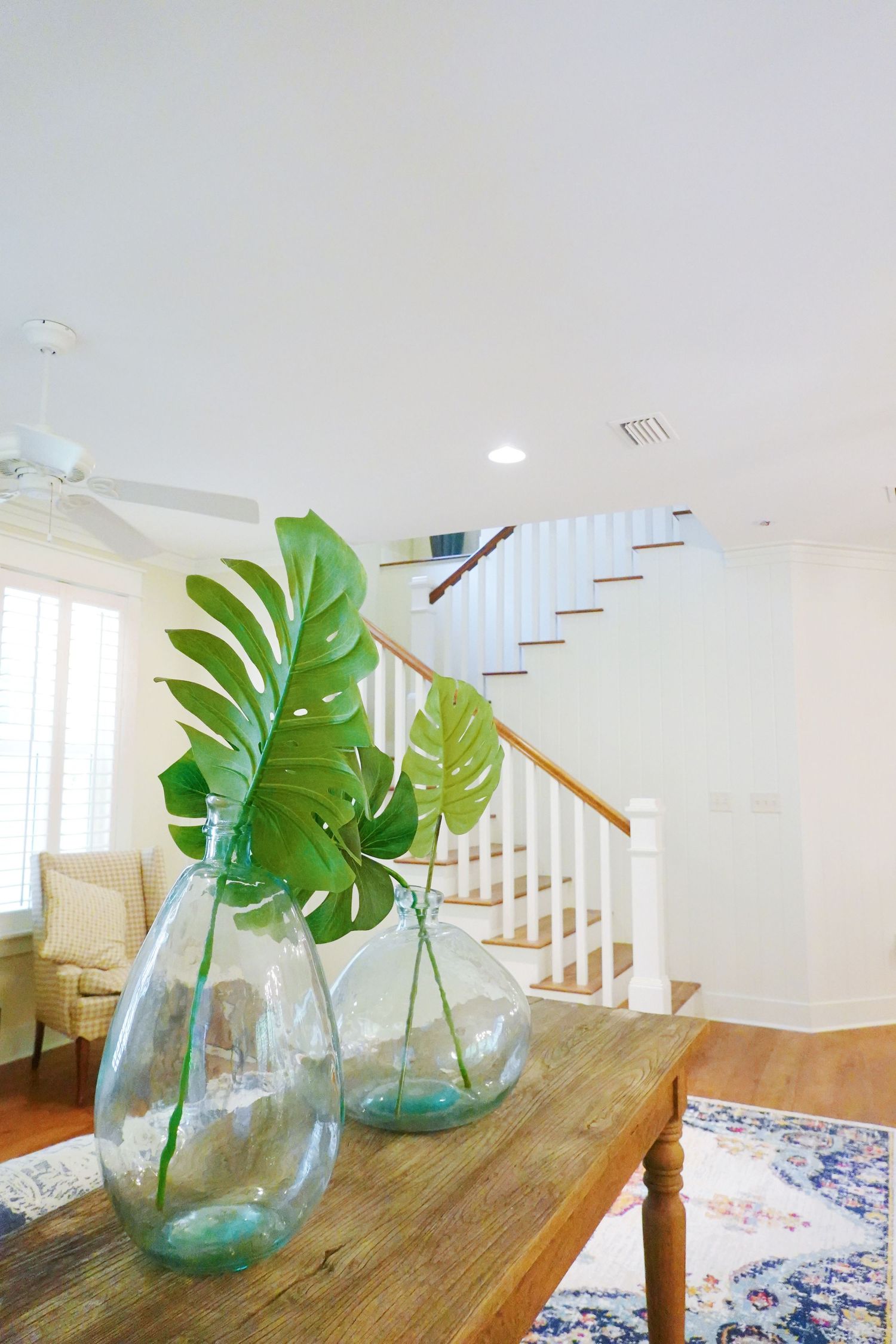Large monstera leaves in glass vases on a wooden table with white staircase and blue rug in background.