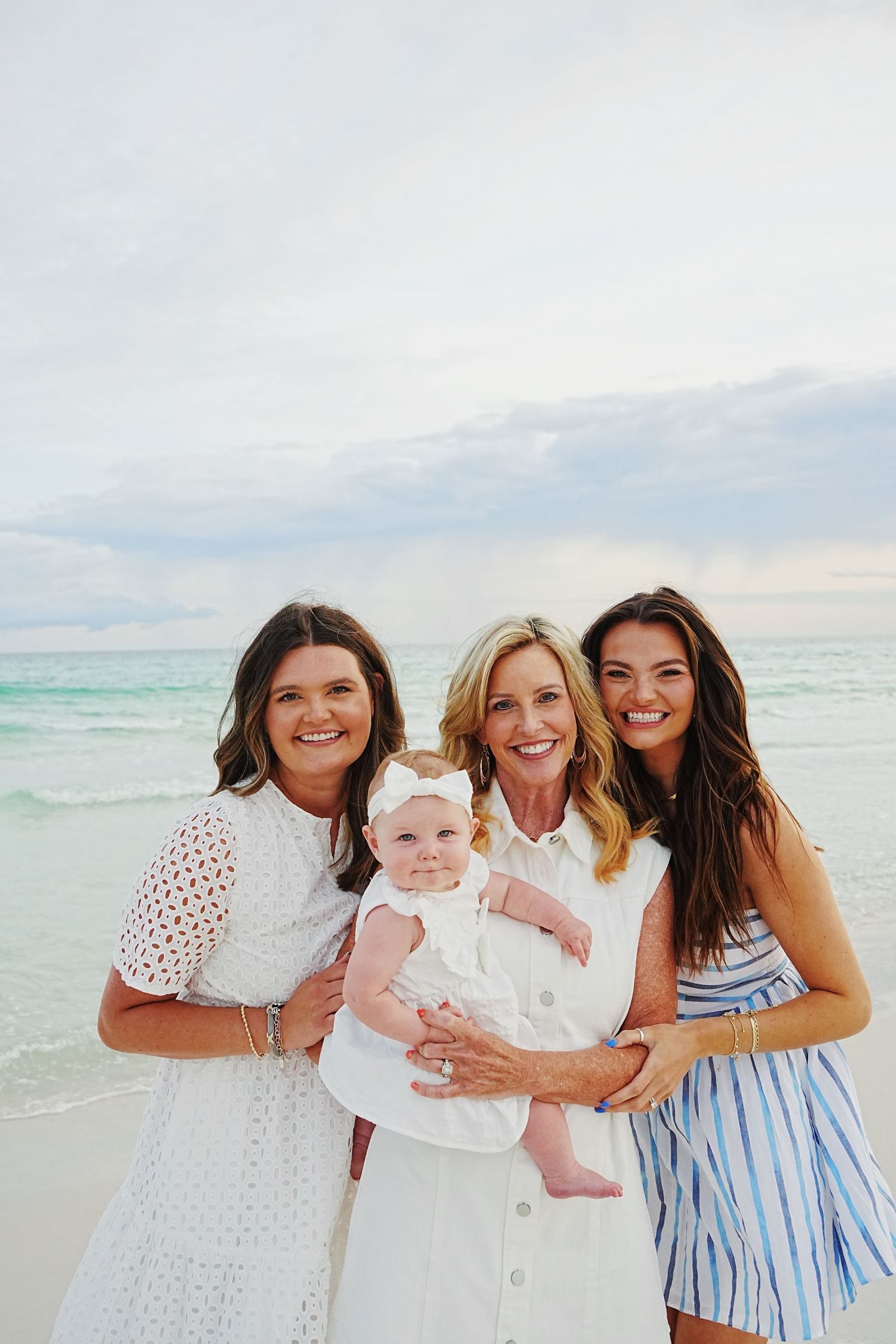 A group of people in white and blue outfits smile together while holding a baby on a sunny beach.
