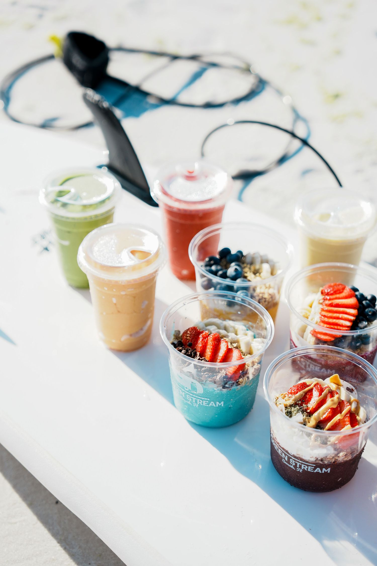 Colorful smoothies and acai bowls topped with fresh berries and fruits arranged on a white surface in bright sunlight.