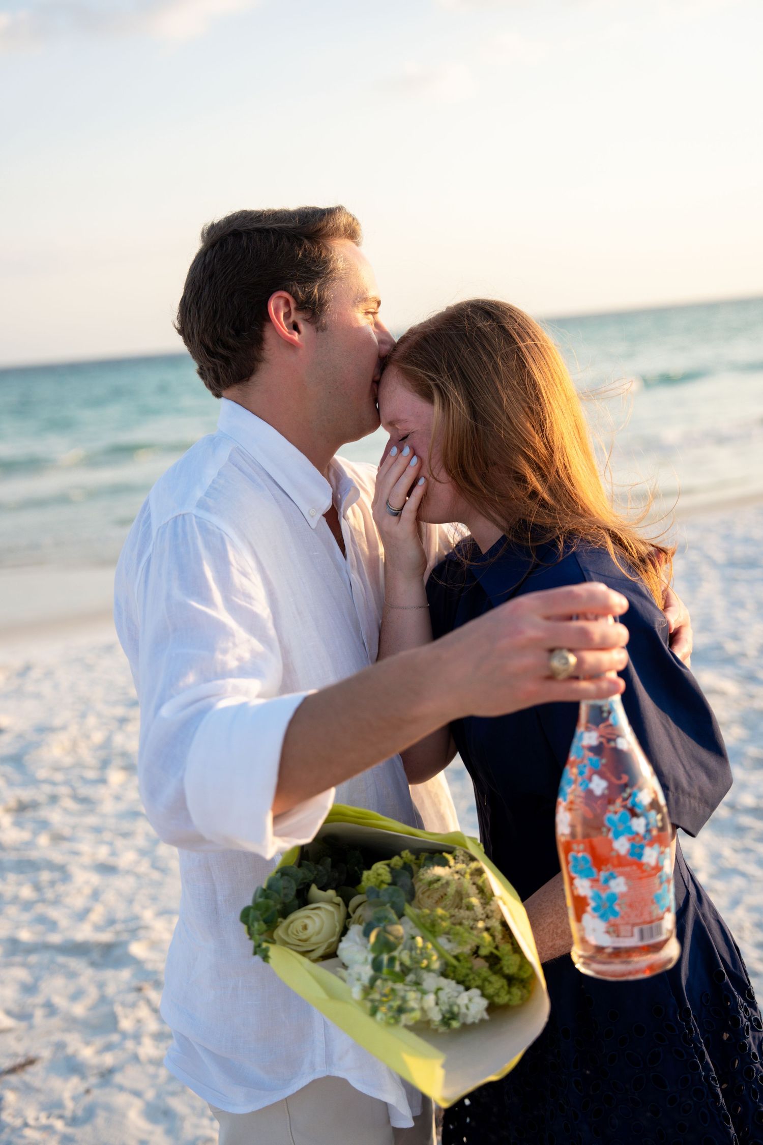 Couple kissing on beach at sunset, man holding flowers and champagne bottle, romantic celebration by the ocean.