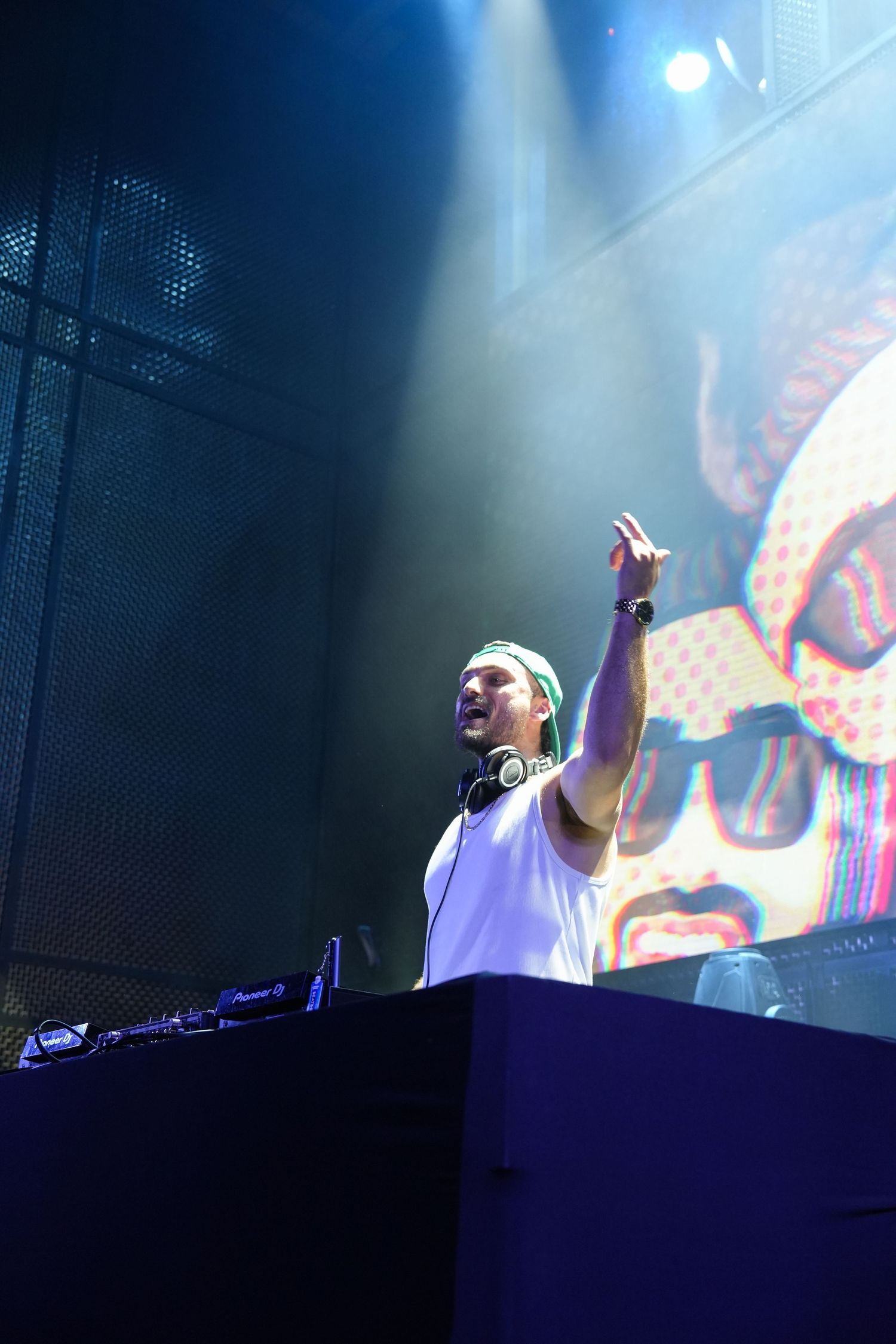 DJ performs behind decks at an electronic music concert with vibrant visual projections in the background.
