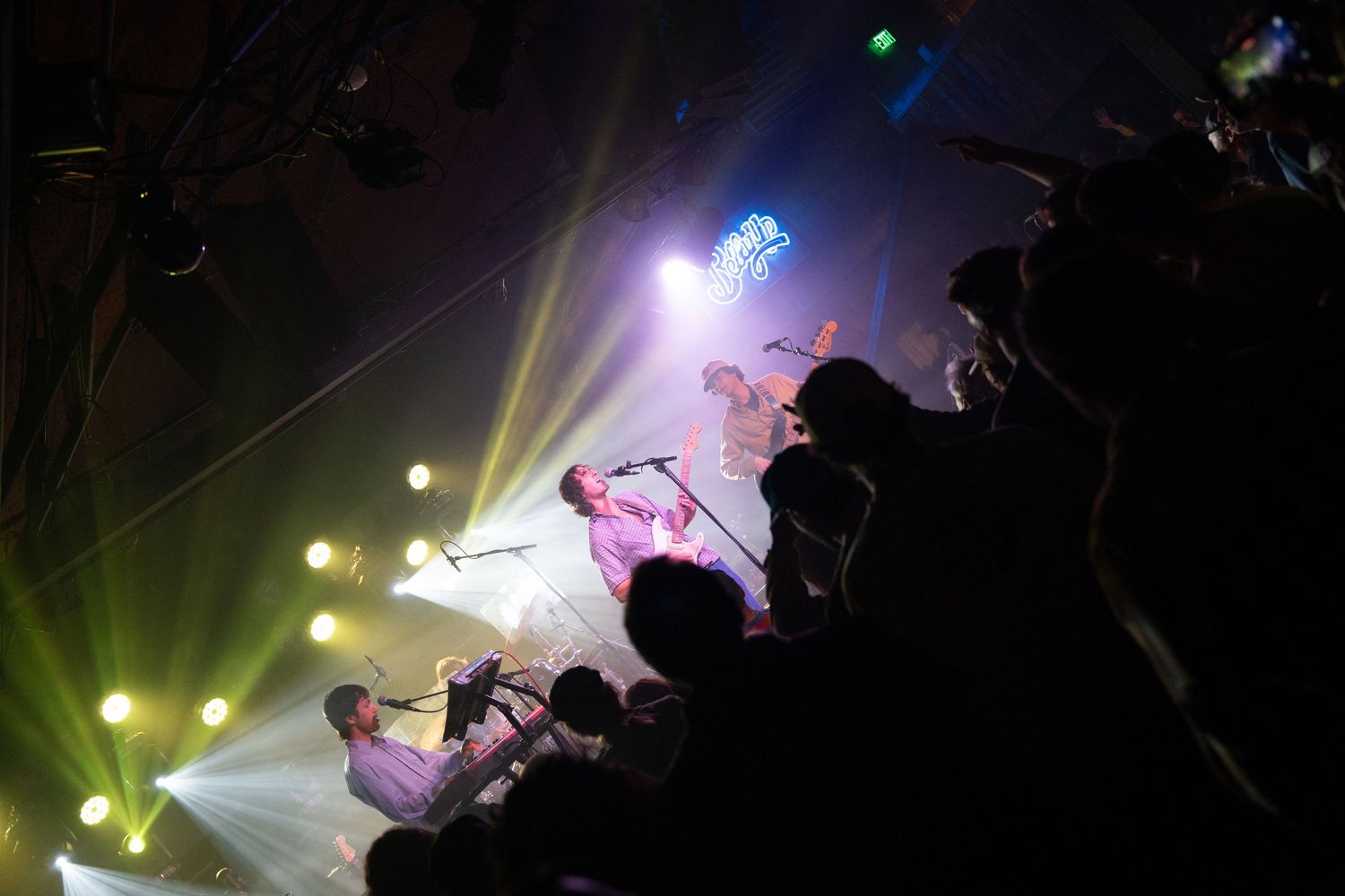Silhouettes of musicians performing on stage with dramatic purple and green lighting effects in the background.