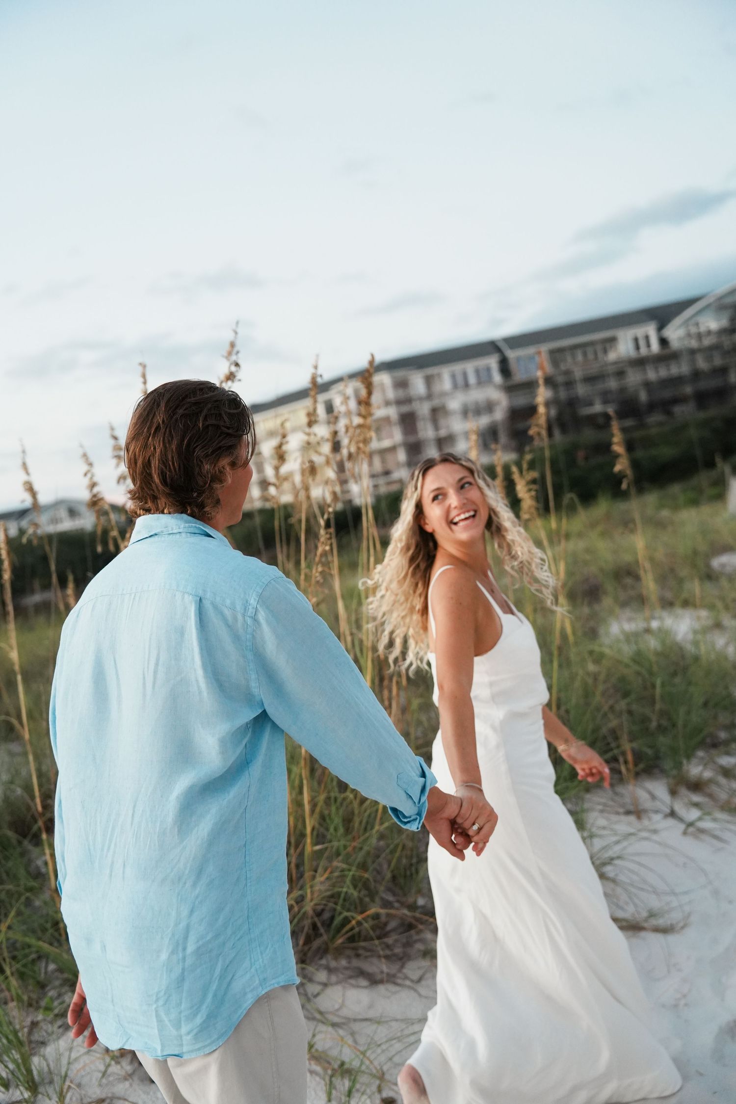 A couple walks hand in hand along a beach path with sea oats swaying in the breeze at sunset.