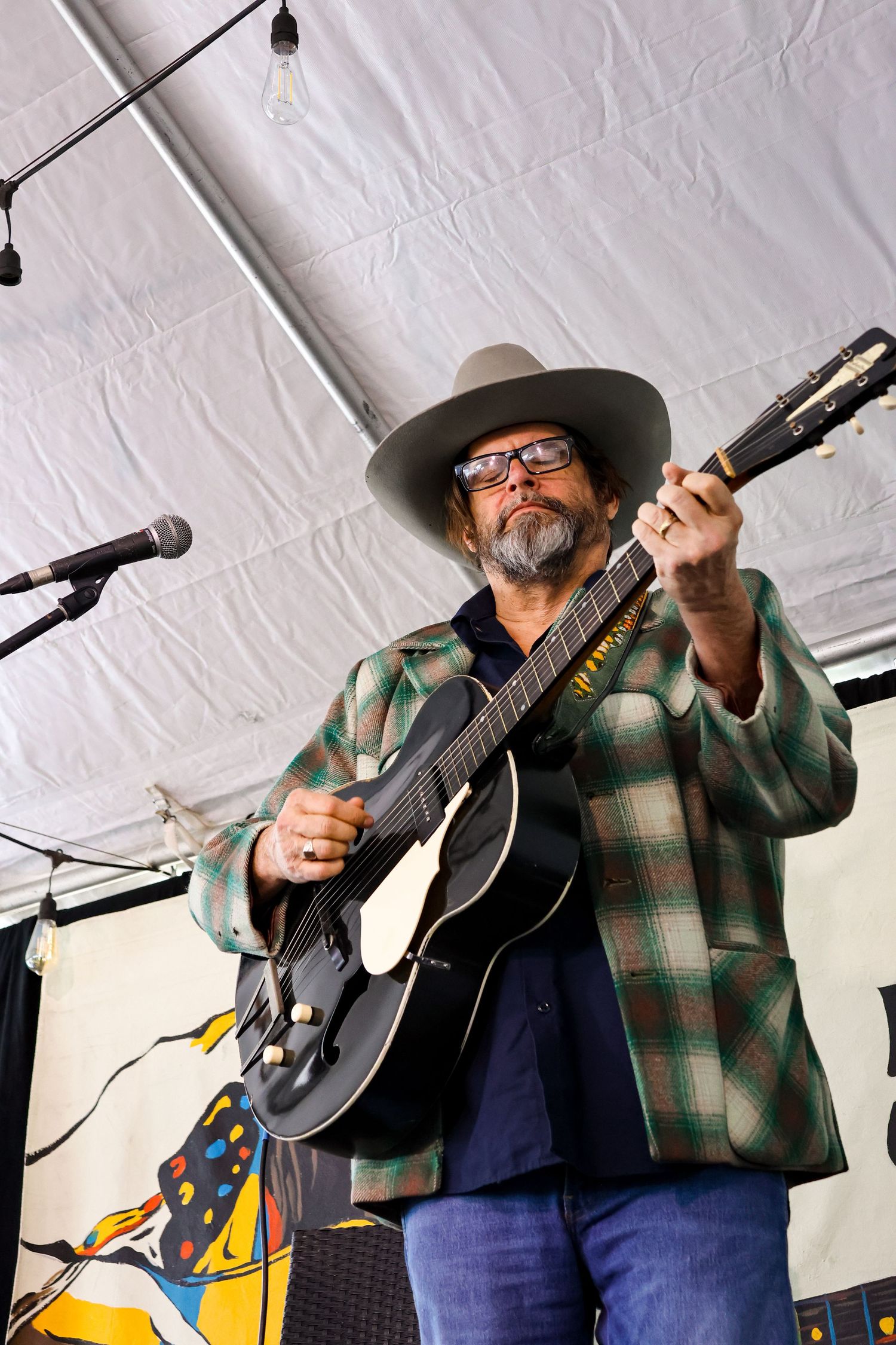 Musician playing acoustic guitar wearing plaid jacket and cowboy hat under tent lighting.