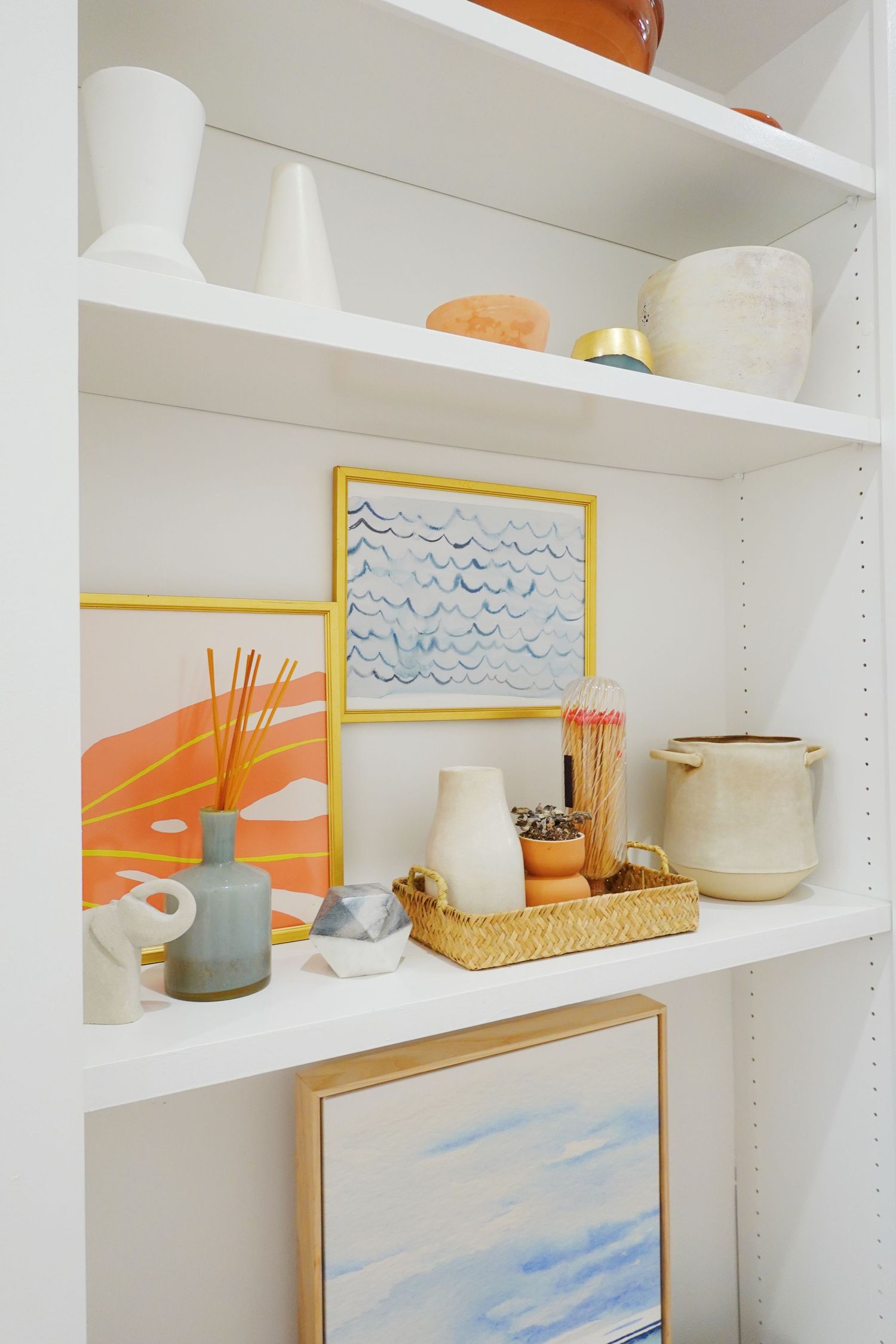 Modern white shelving unit displaying coastal decor, artwork, and ceramic vessels.