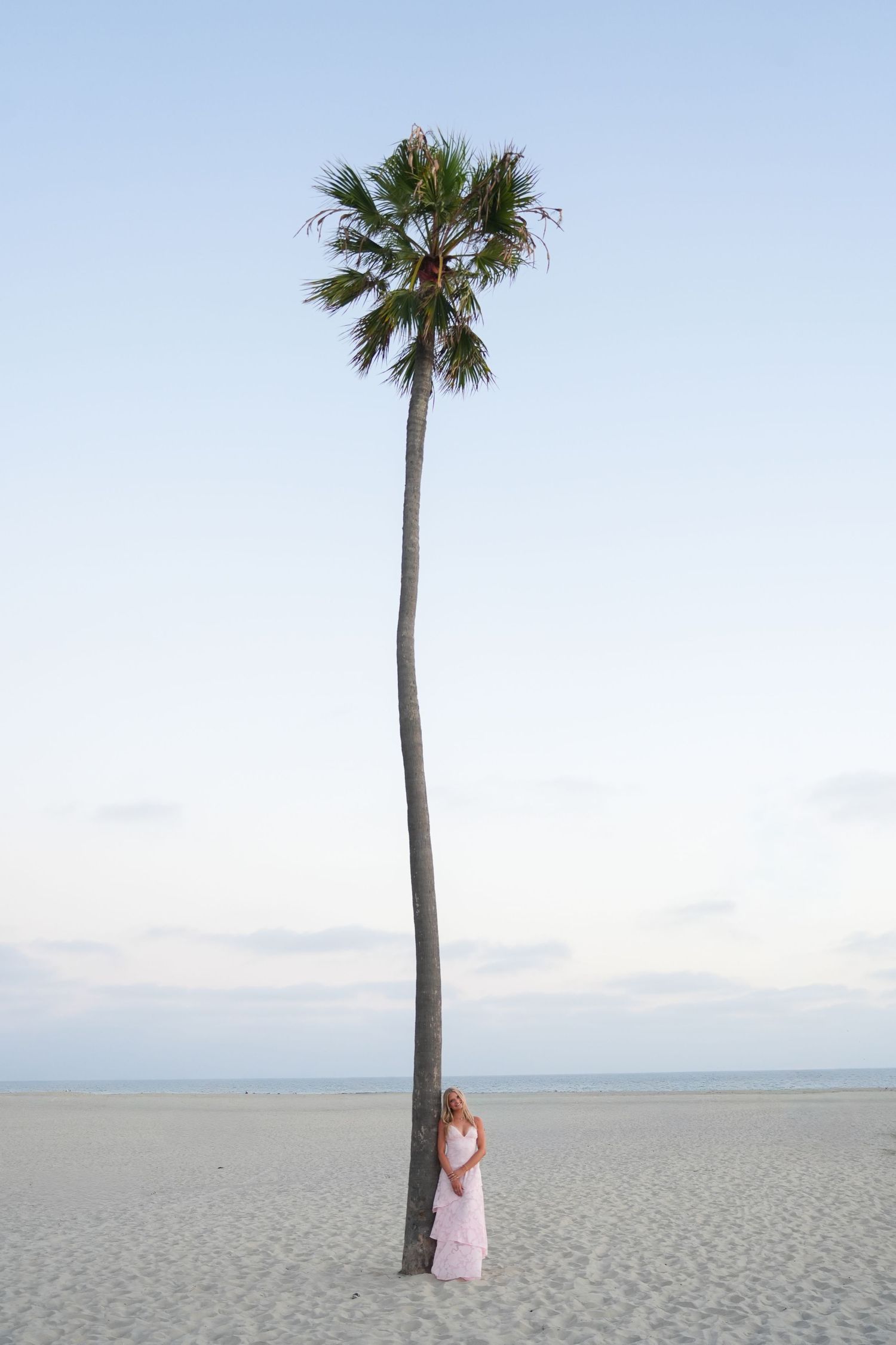 Woman in pink dress standing beside a tall palm tree on a sandy beach at dusk.
