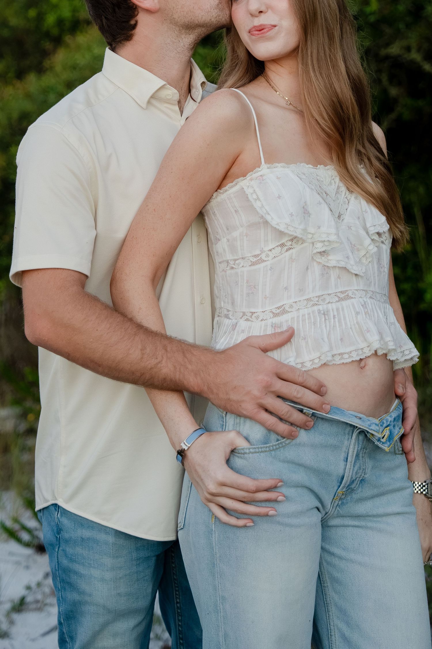 Couple embracing outdoors, woman in white ruffled crop top and jeans, man in beige shirt, romantic pose.