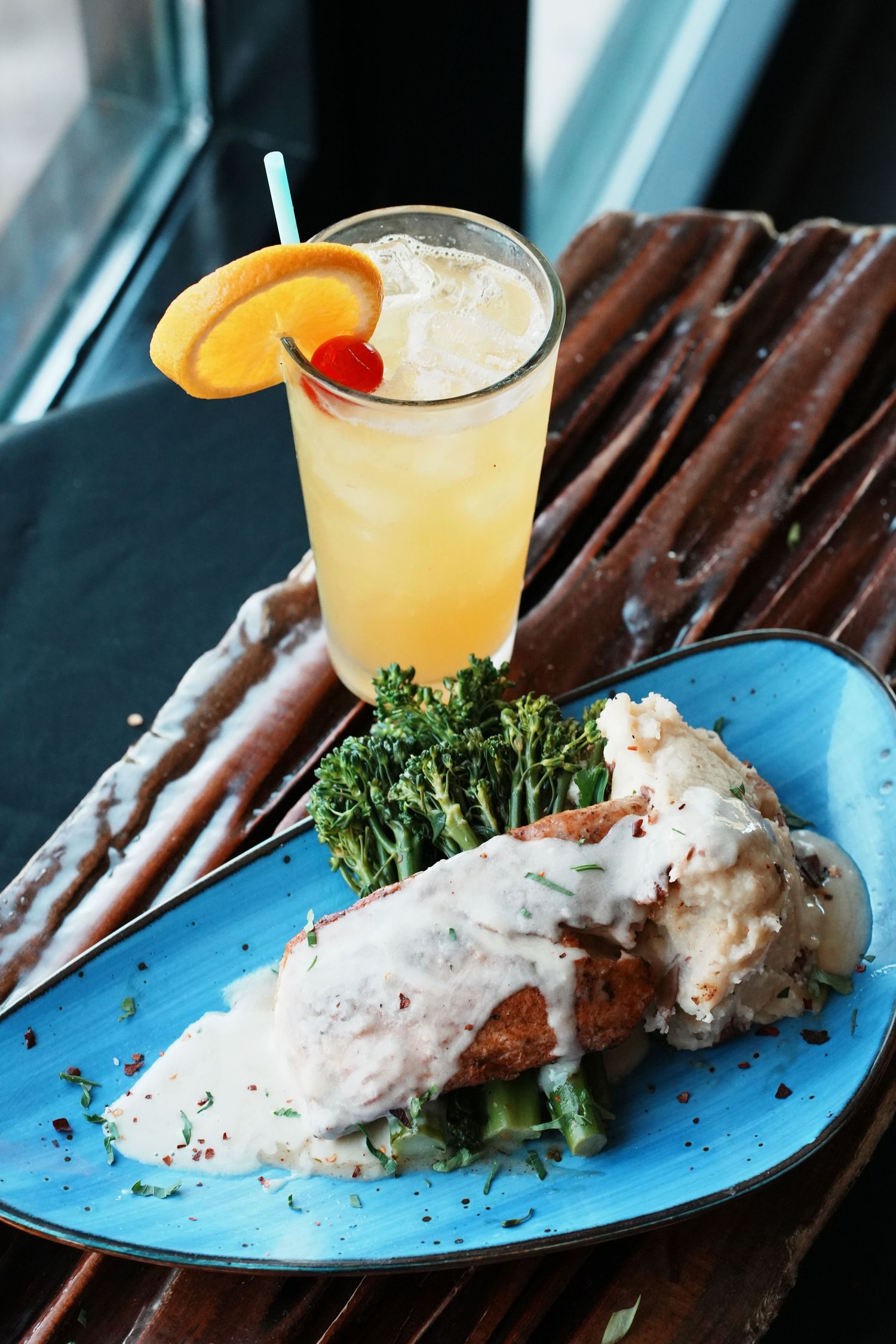 Grilled meat with creamy sauce and broccoli served with a citrus cocktail on a blue plate.