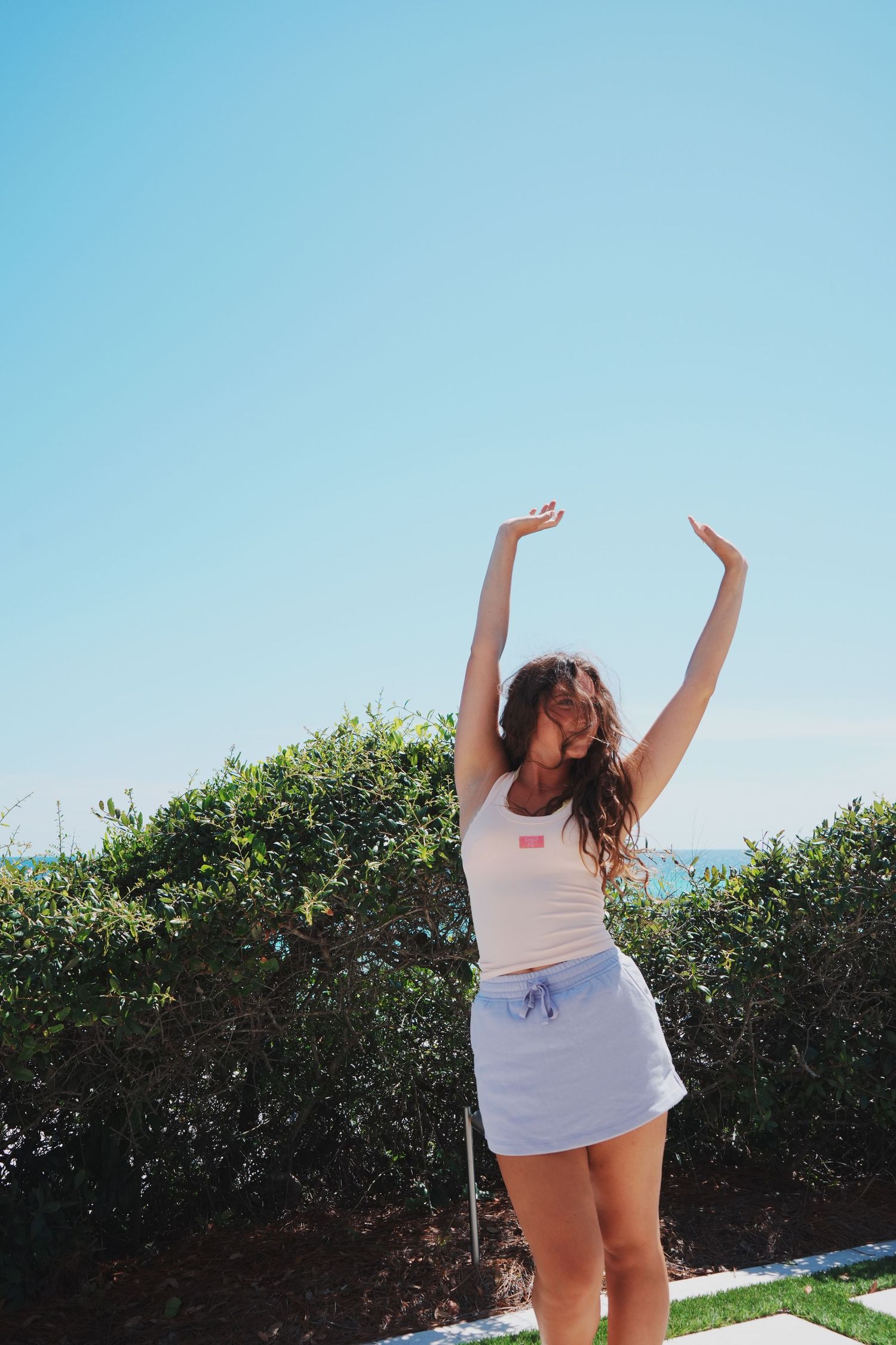 Person in summer attire dancing with arms raised against blue sky and greenery.