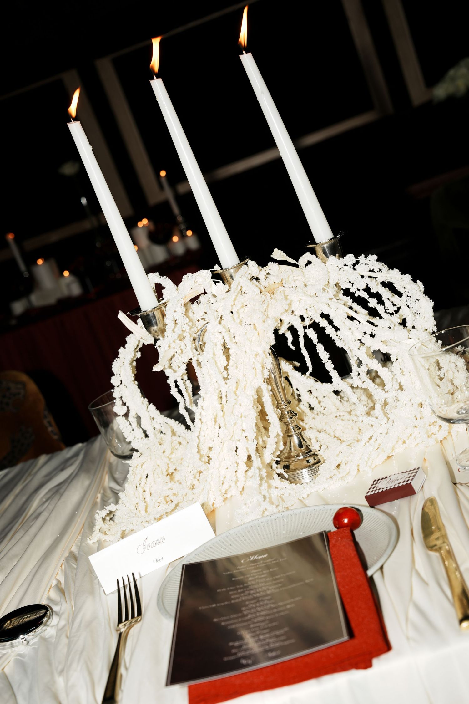 Elegant wedding table setting with lit white taper candles, floral centerpiece, red menu frame, and place card.