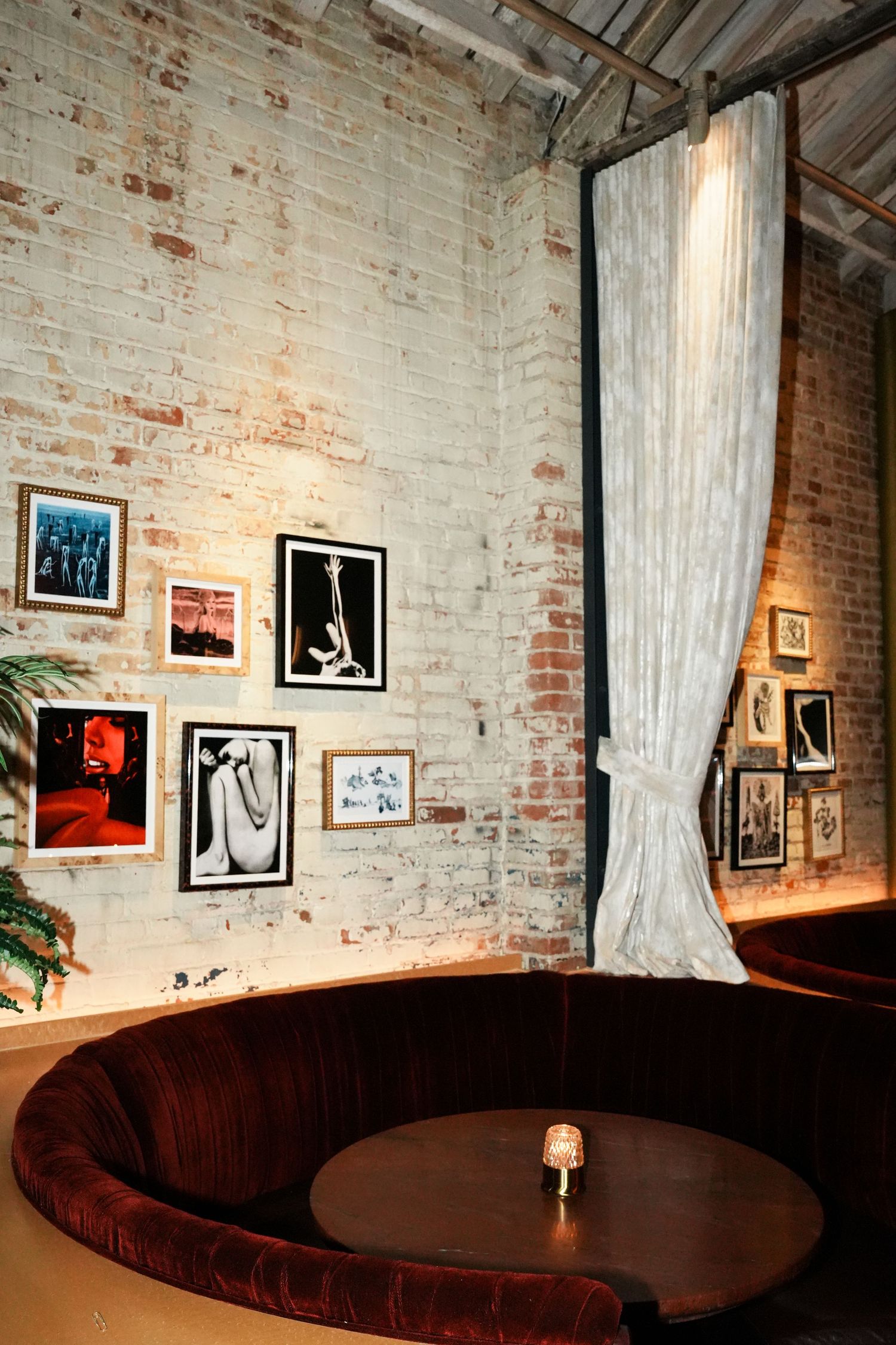 Cozy restaurant booth with curved velvet seating, round table, and art gallery wall in brick interior.