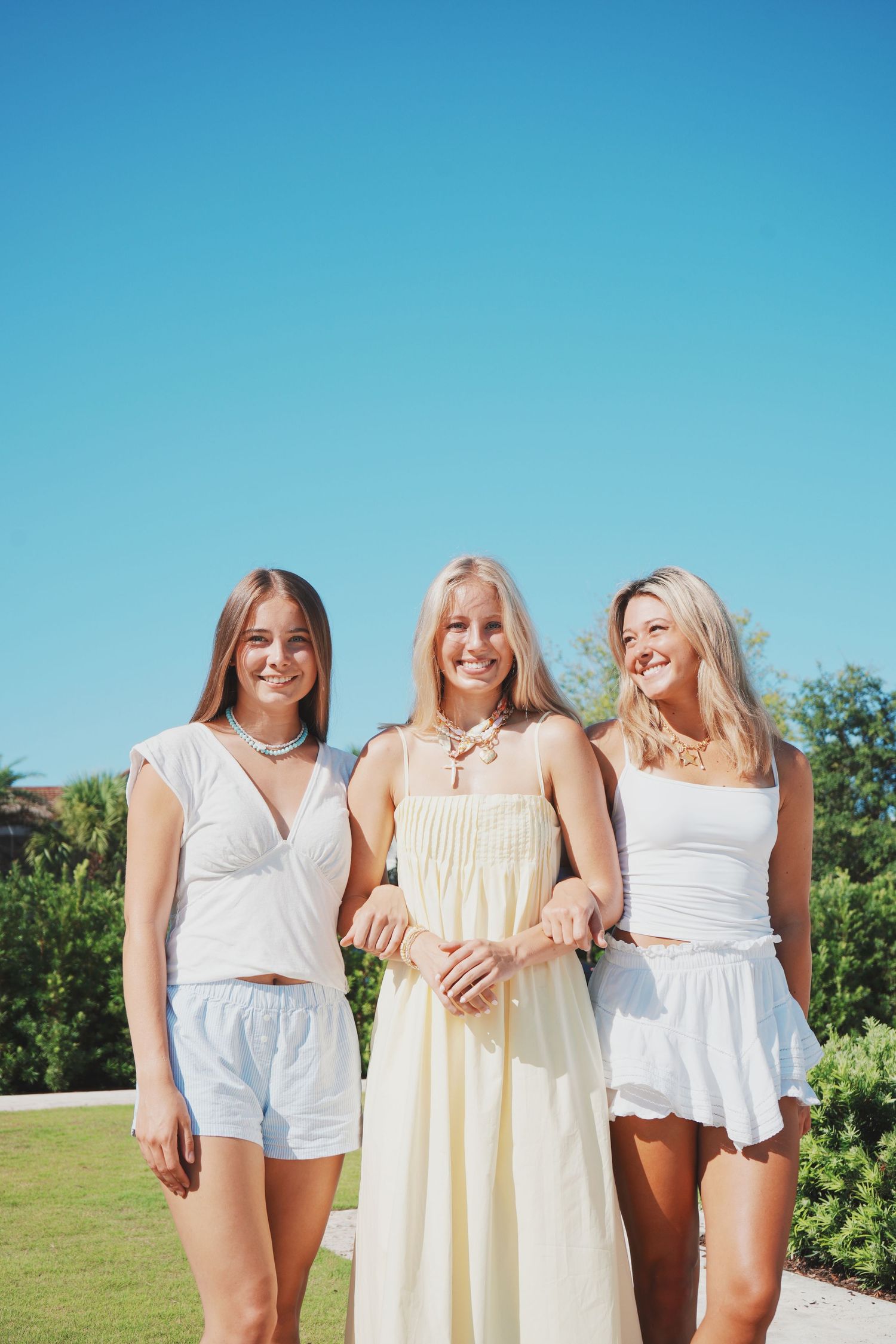 Three women in white and yellow summer outfits enjoying a sunny day outdoors against a bright blue sky.