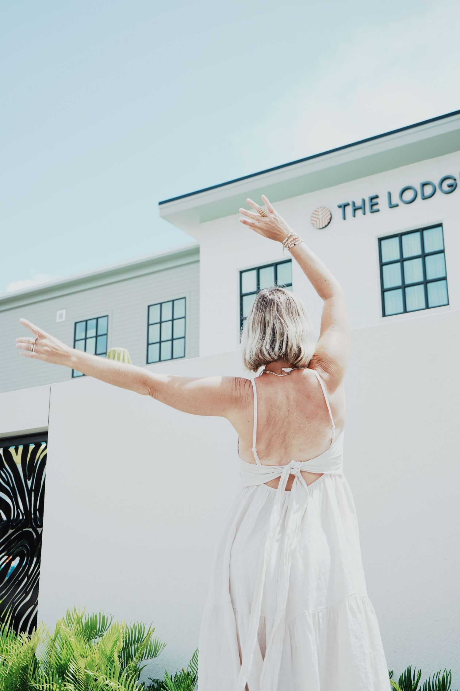 Person in flowing white dress strikes graceful dance poses against modern white building with palm trees and windows.