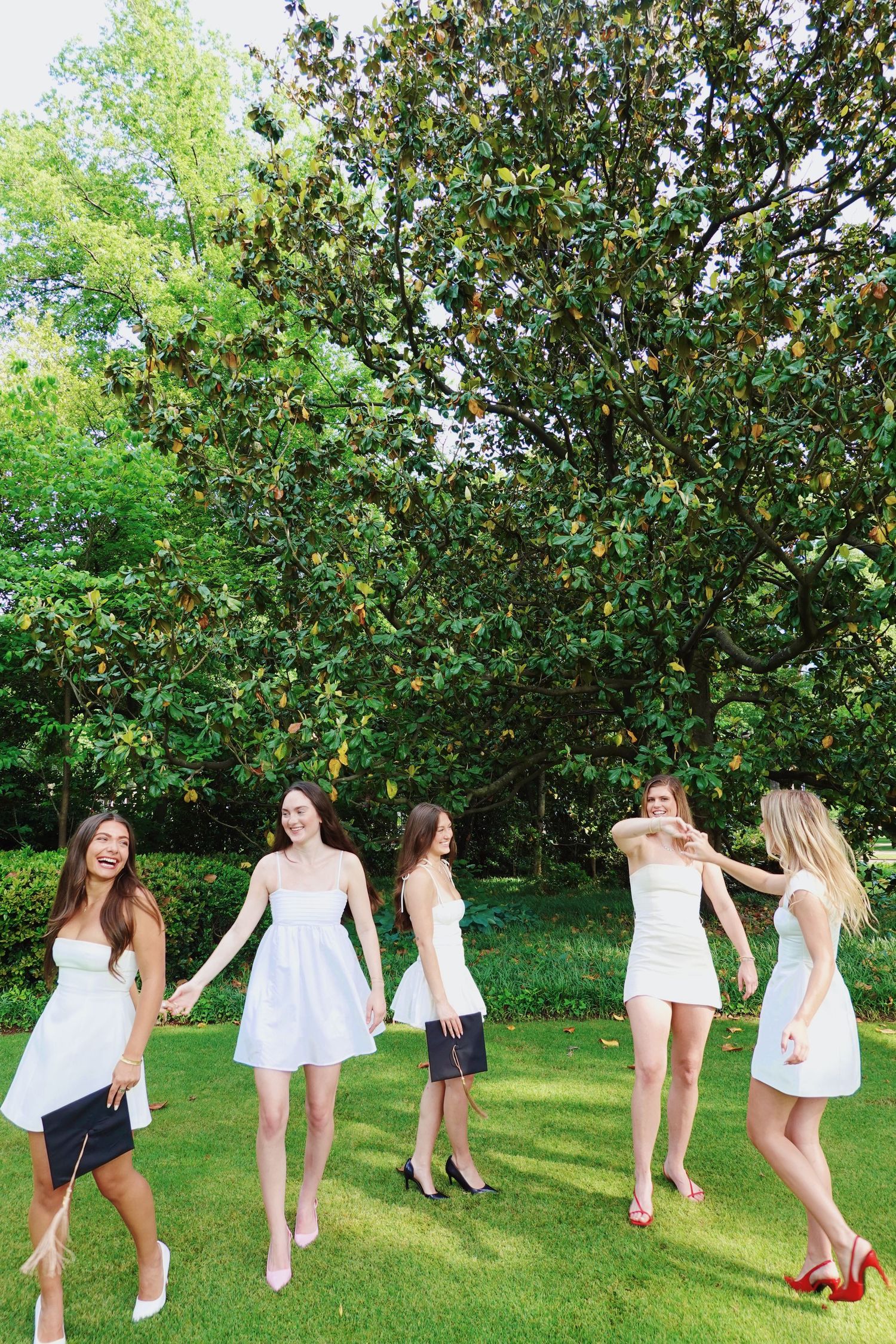 Friends in white dresses playfully posing with graduation caps under magnolia trees.